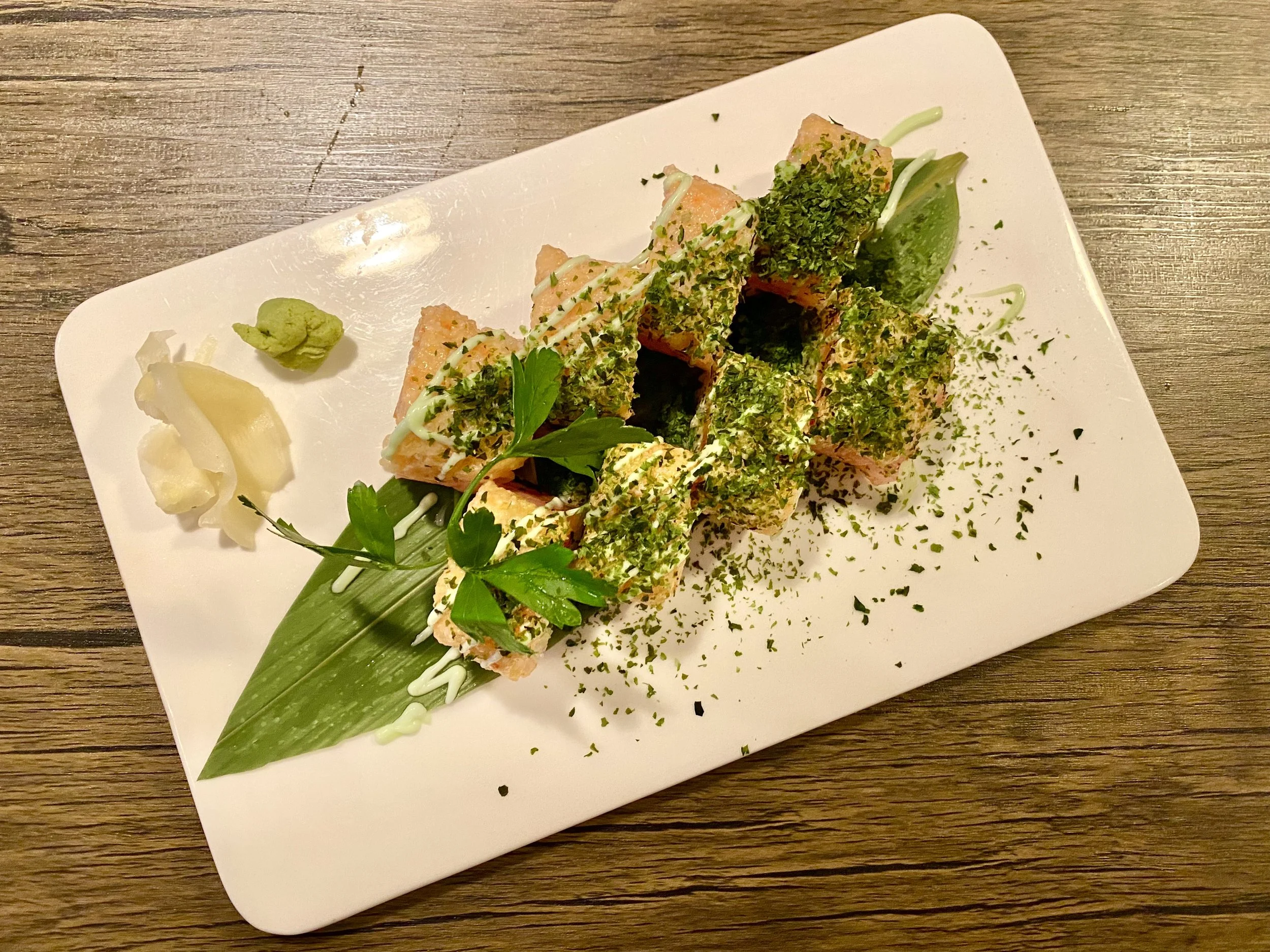 A plate of sushi rolls topped with green herbs and drizzled with sauce, served with wasabi, pickled ginger, and a lime wedge on a wooden table.