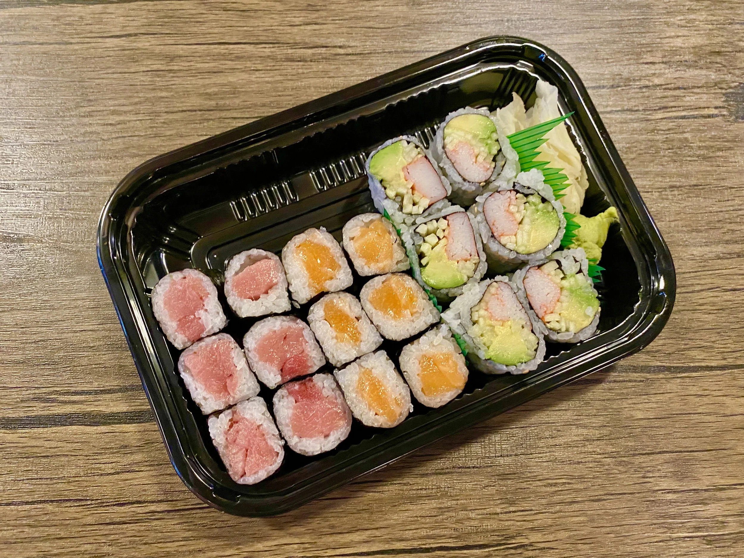 Sushi rolls arranged in a black plastic container on a wooden surface, featuring varieties with pink, orange, and green fillings.