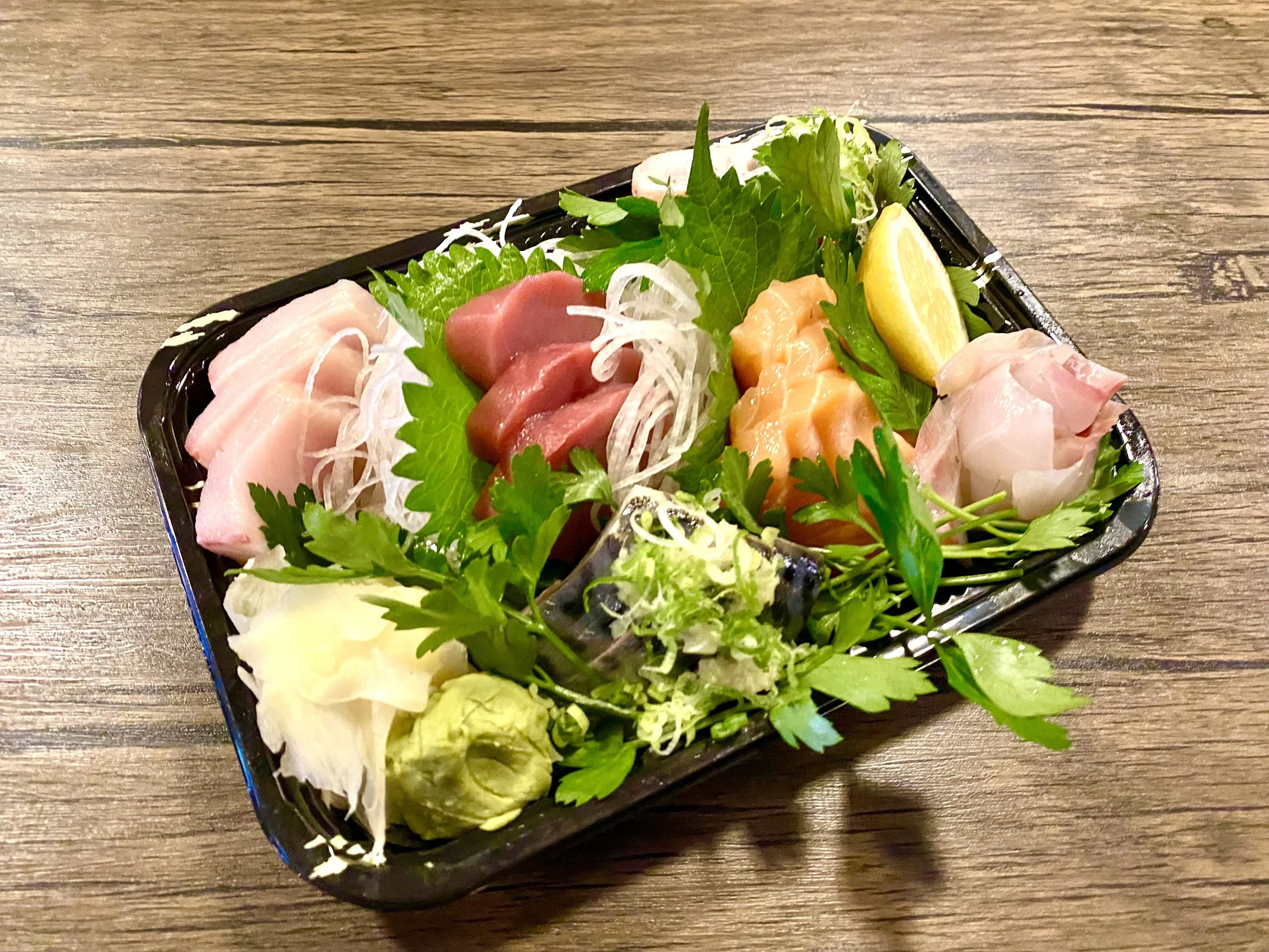 Assorted sashimi with slices of raw fish, garnished with green herbs, lemon wedge, and wasabi on a black tray.