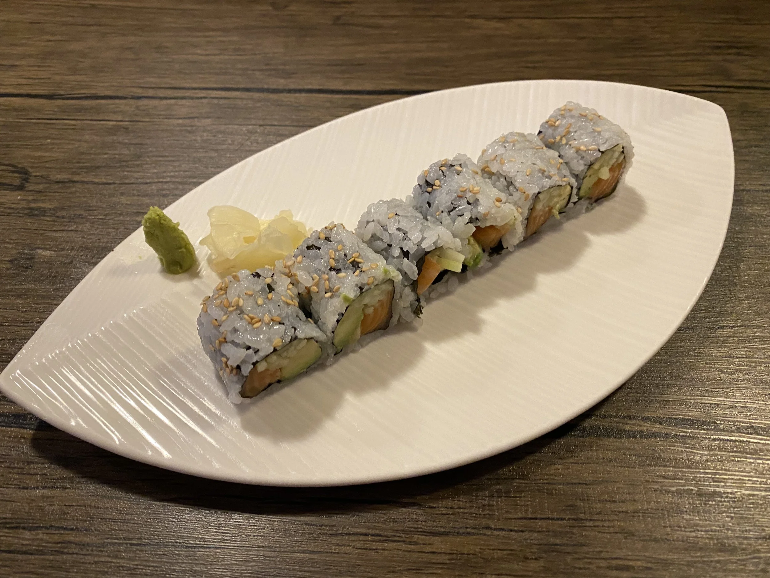 Sushi rolls with sesame seeds on a white oval plate, accompanied by wasabi and pickled ginger on a wooden table.