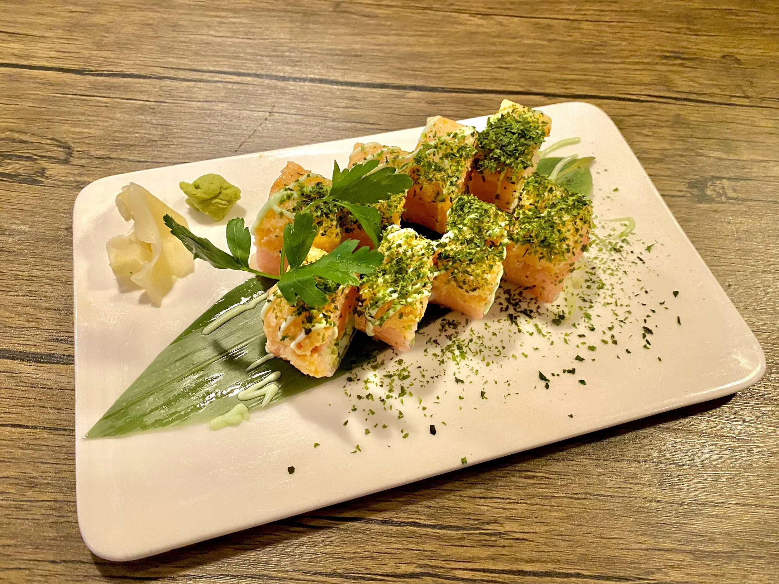 Sushi roll topped with green herbs, garnished with a sprig of parsley, served on a white rectangular plate with wasabi, pickled ginger, and decorative green and white sauce drizzles, placed on a wooden table.