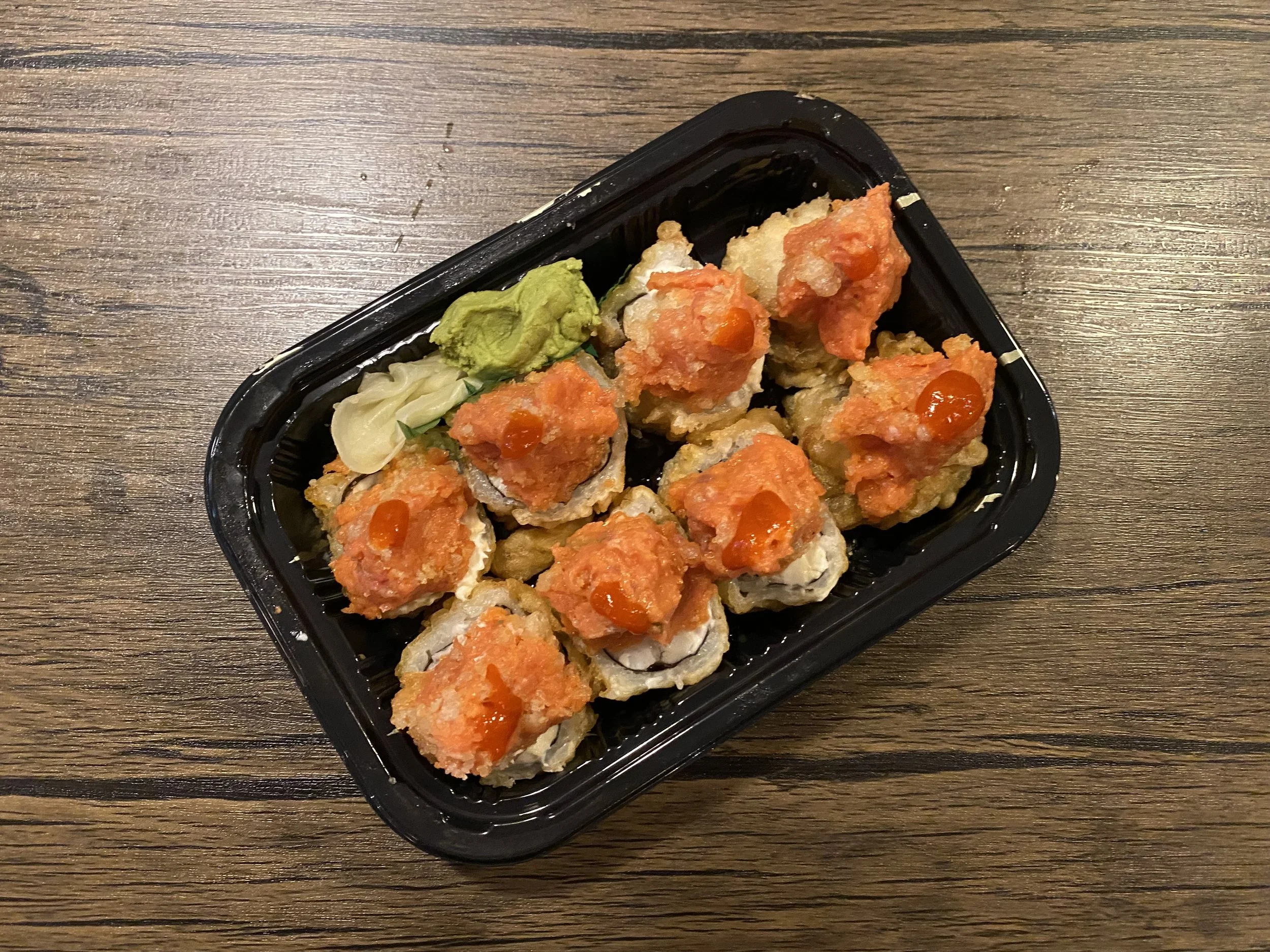 A black plastic tray containing sushi rolls topped with orange fish roe and drizzled with sauce, garnished with wasabi and pickled ginger, on a wooden table.