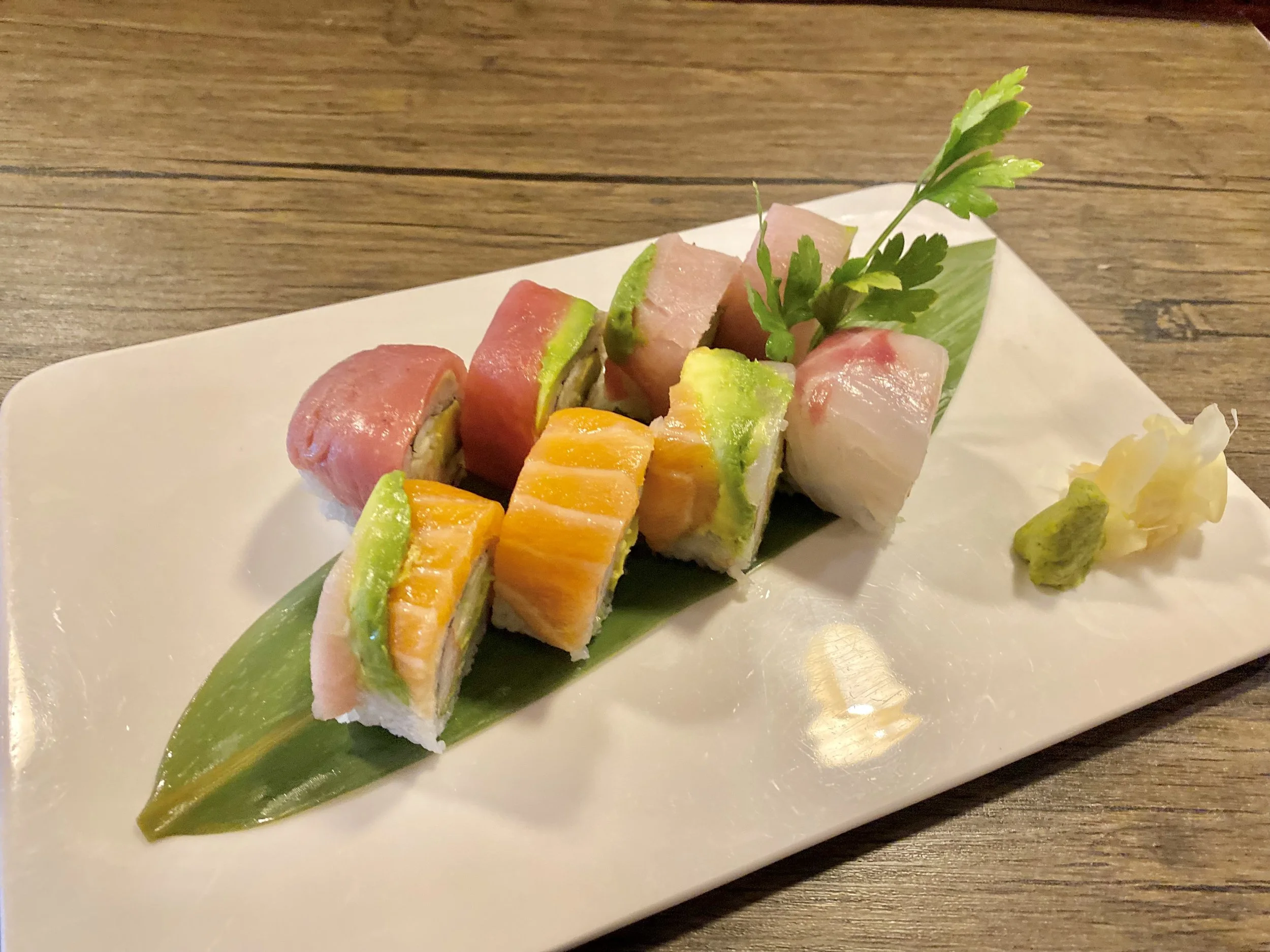 Assorted sushi rolls arranged on a white plate with a green leaf, garnished with parsley, wasabi, and pickled ginger.