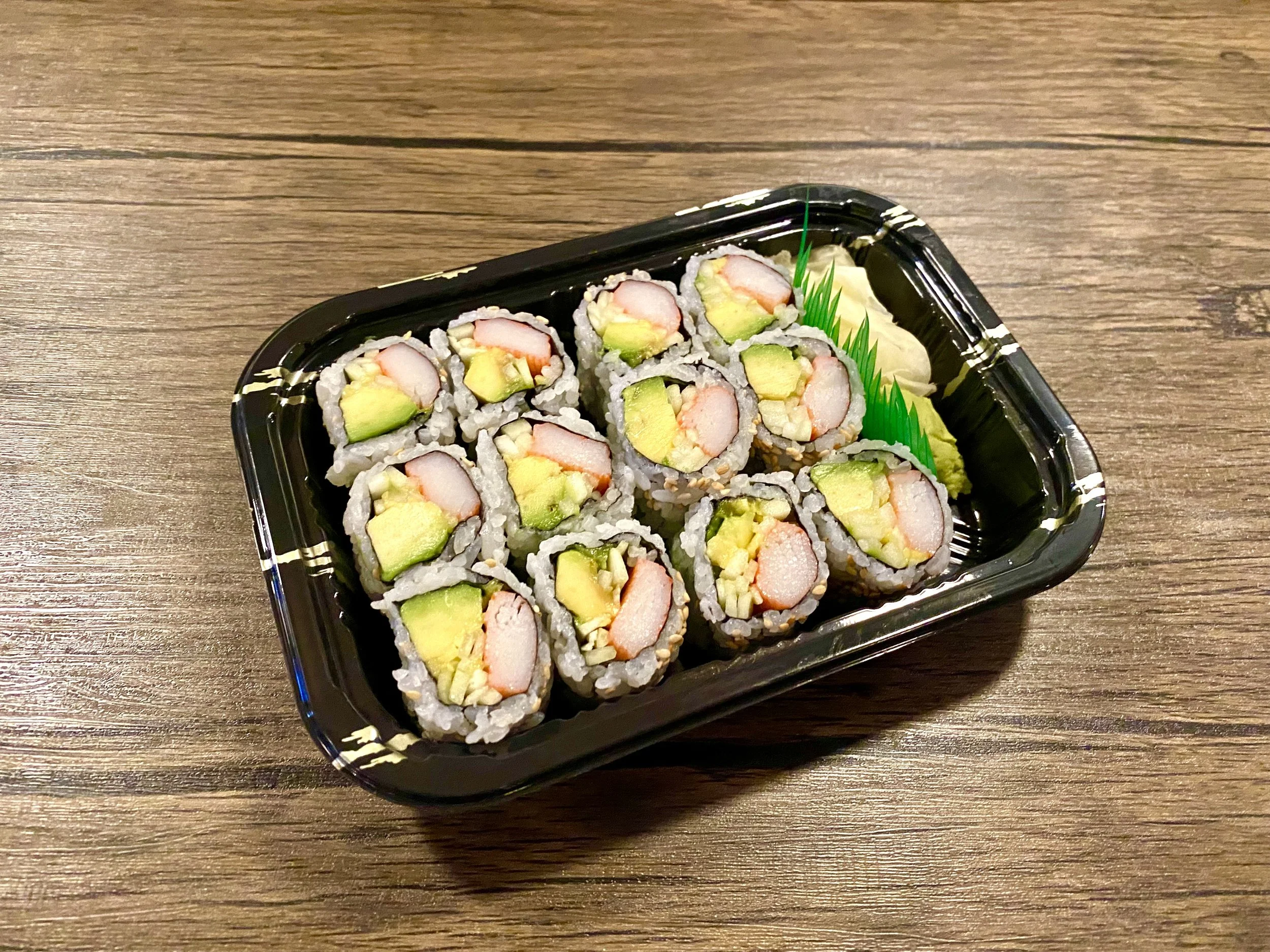 Sushi rolls with crab, avocado, and cucumber in a black plastic tray on a wooden table.