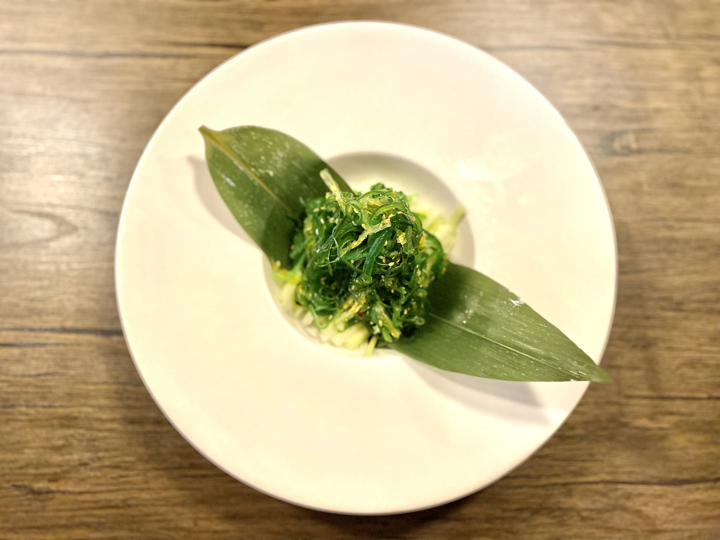 A plate with a green salad garnished with two large green leaves arranged diagonally, on a wooden table.
