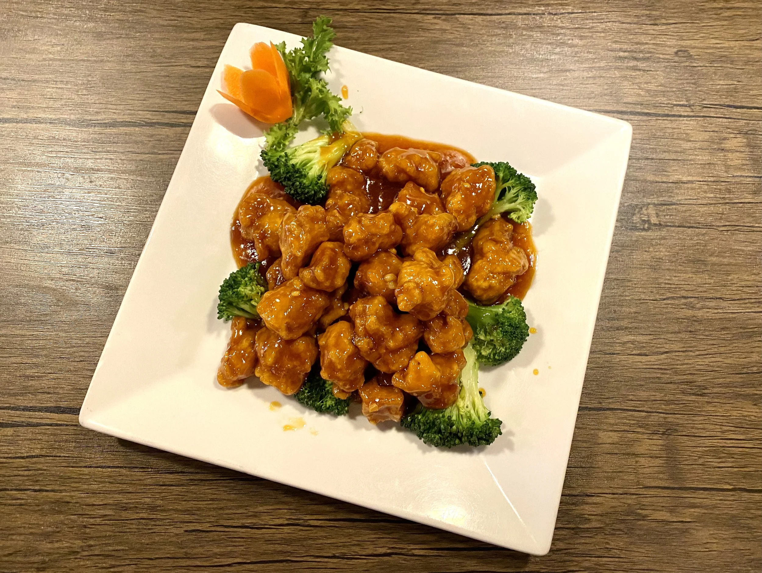 A white square plate with crispy chicken pieces coated in orange sauce, garnished with broccoli florets and a decorative carrot flower. The plate is on a wooden table.