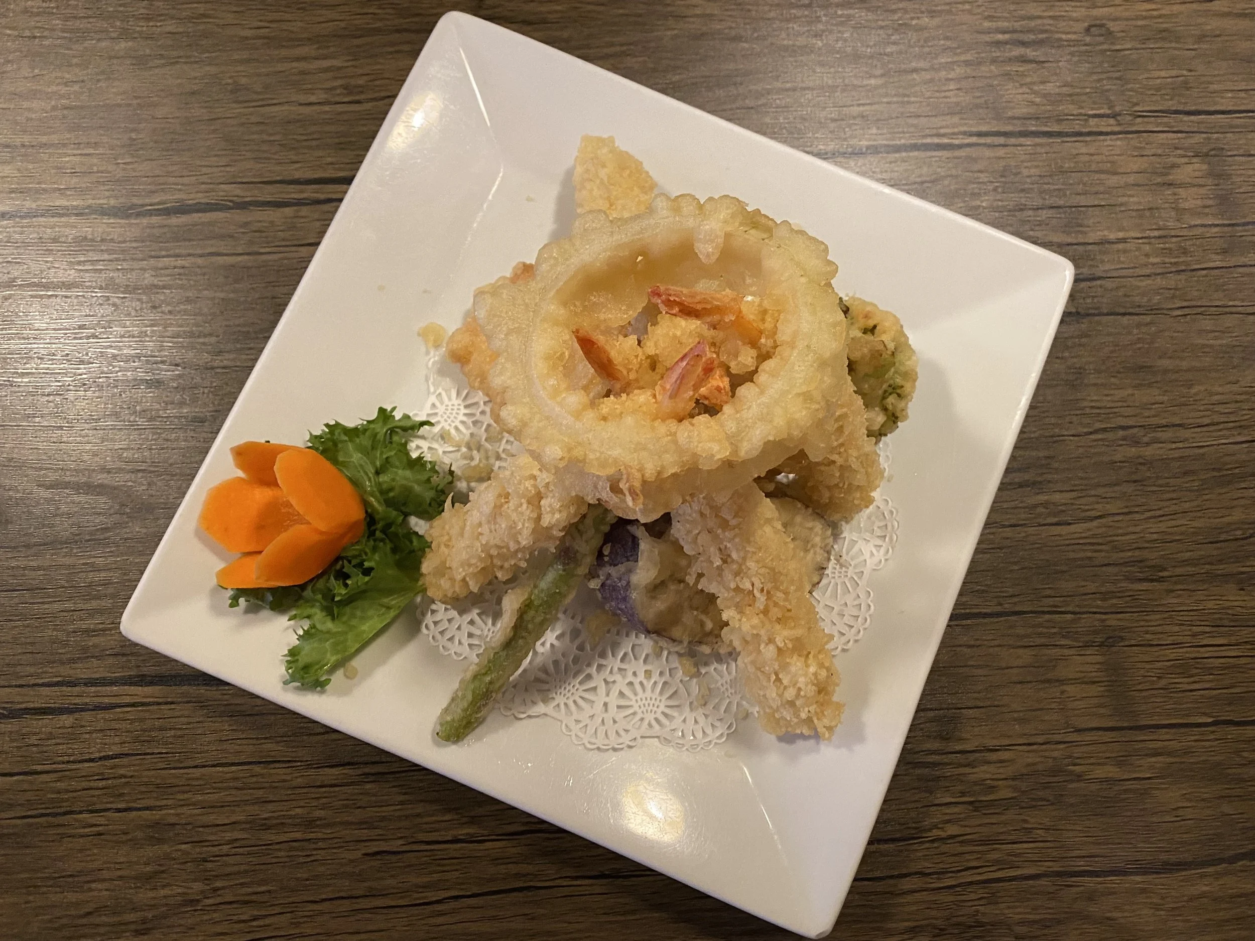 A white square plate with fried calamari rings and vegetable tempura, garnished with a small lettuce leaf and an orange edible flower, on a wooden table.