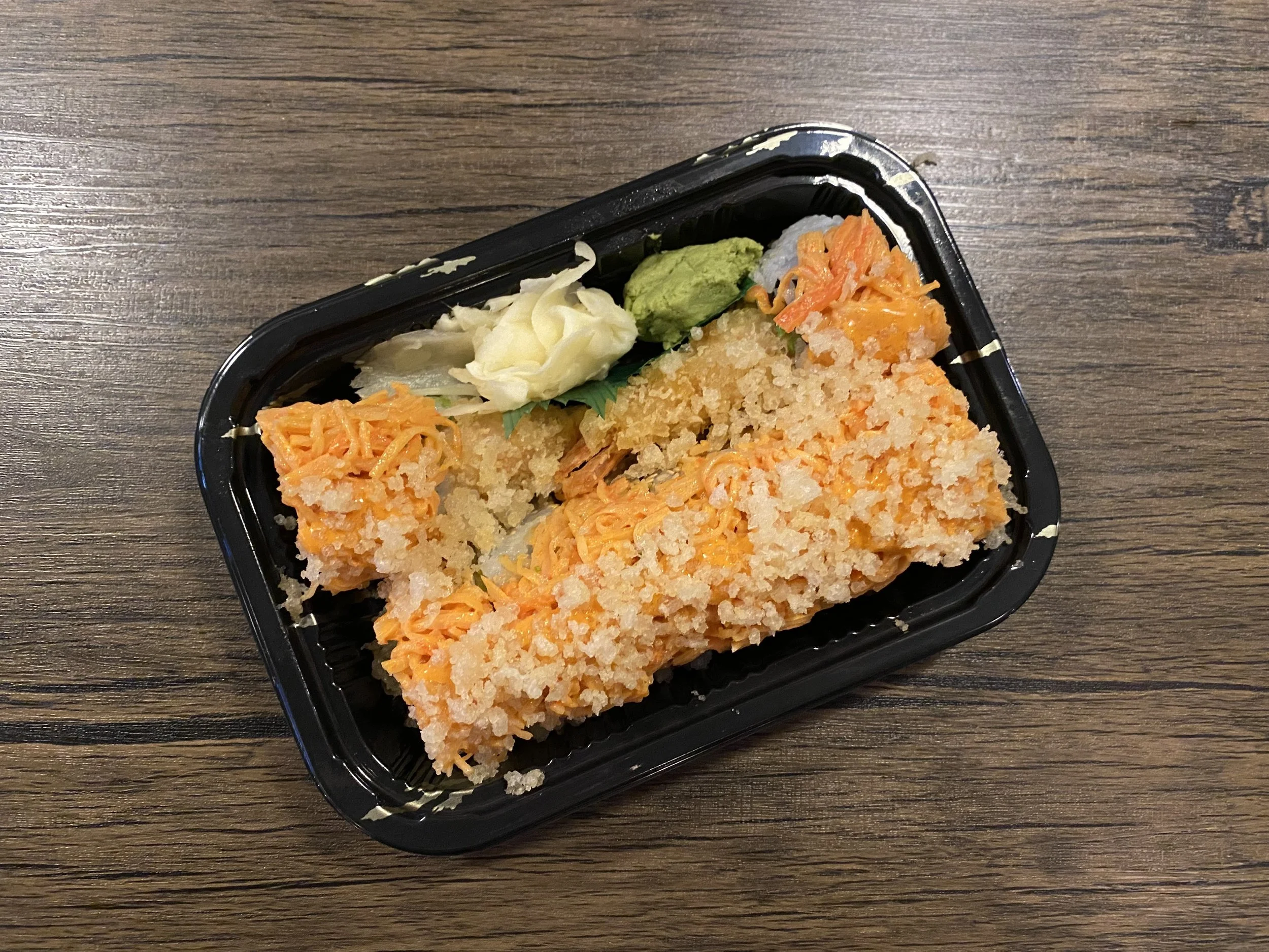 A black plastic container with breaded fried fish fillet, grated carrots, shredded cabbage, wasabi, pickled ginger, and a small portion of rice on a wooden surface.