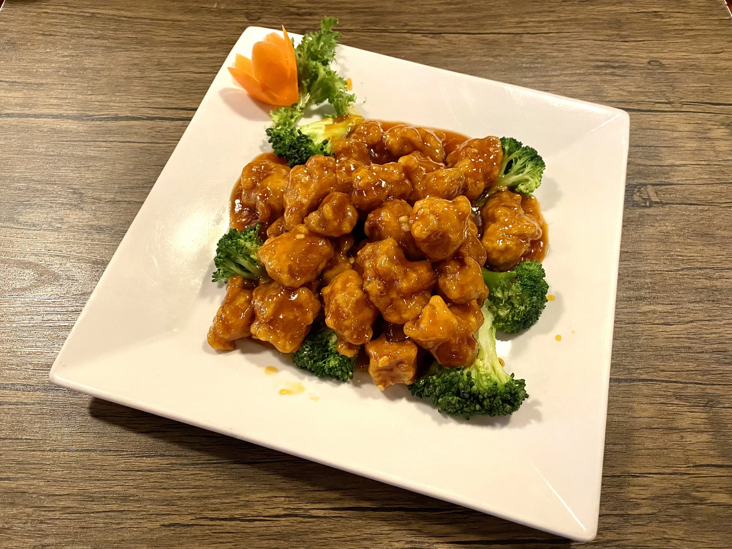 A plate of sweet and sour chicken with broccoli and a decorative carved carrot piece on a wooden table.