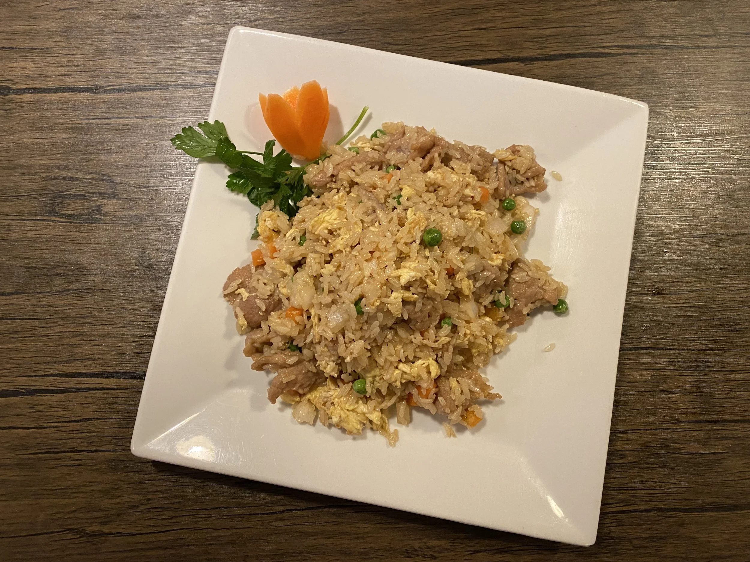 Plate of fried rice with peas, scrambled eggs, small pieces of meat, garnished with a carved carrot flower and green herbs, on a wooden table.