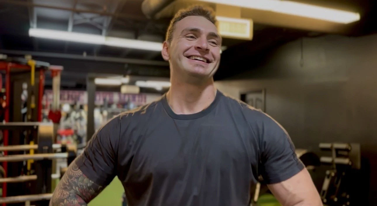 A man smiling, wearing a black athletic shirt, in a gym with workout equipment in the background.
