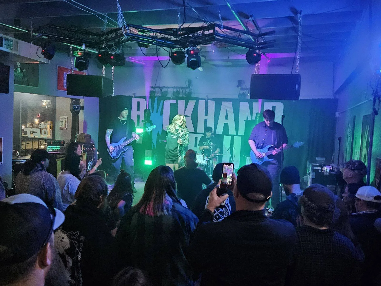 A band performs on stage at a live music event with an audience watching. The stage has a backdrop that reads 'BREAKFAST' and colorful lighting.