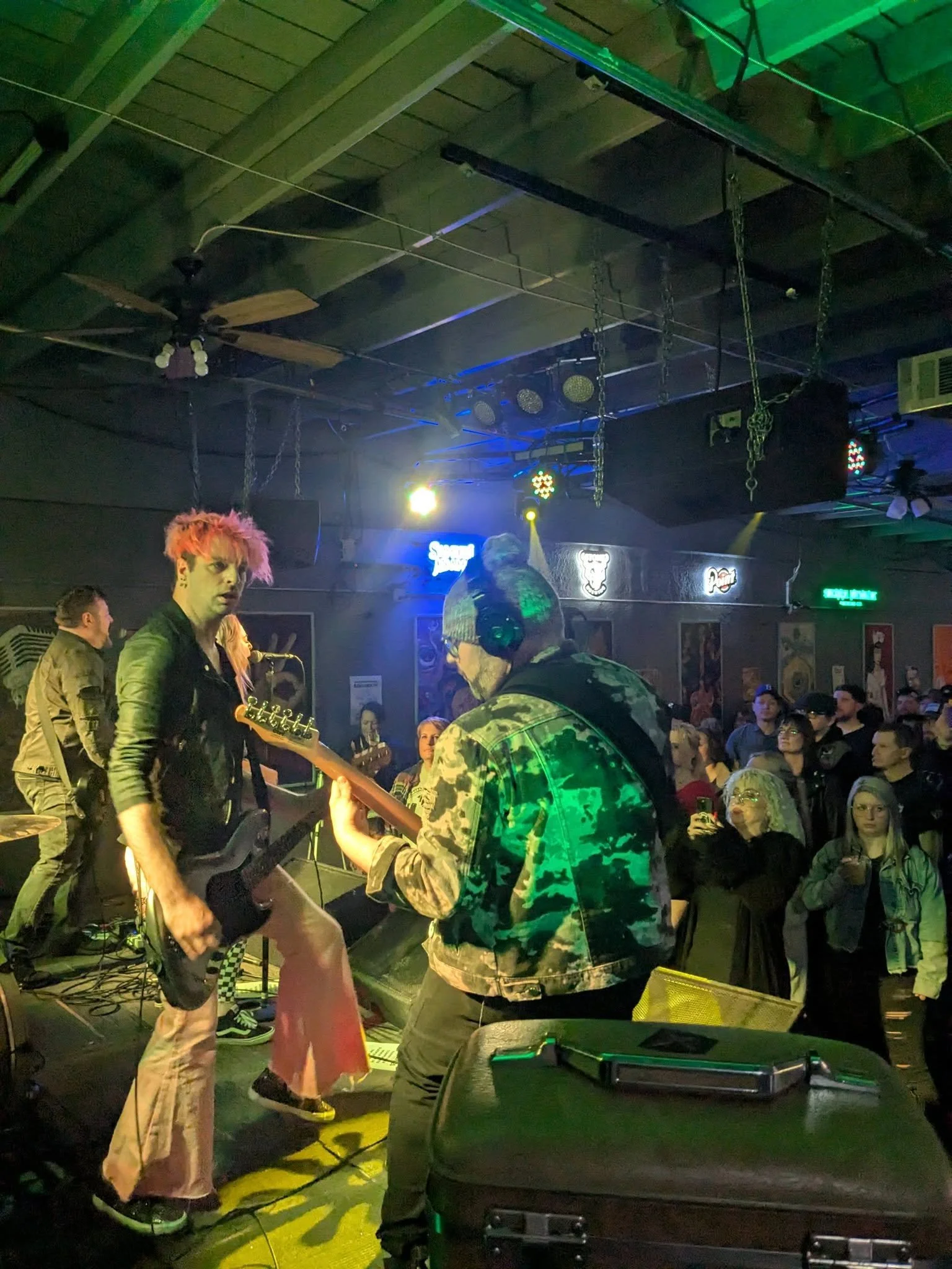 People performing live music on a stage in a nightclub or concert venue, with audience watching, colorful stage lights, and music equipment.