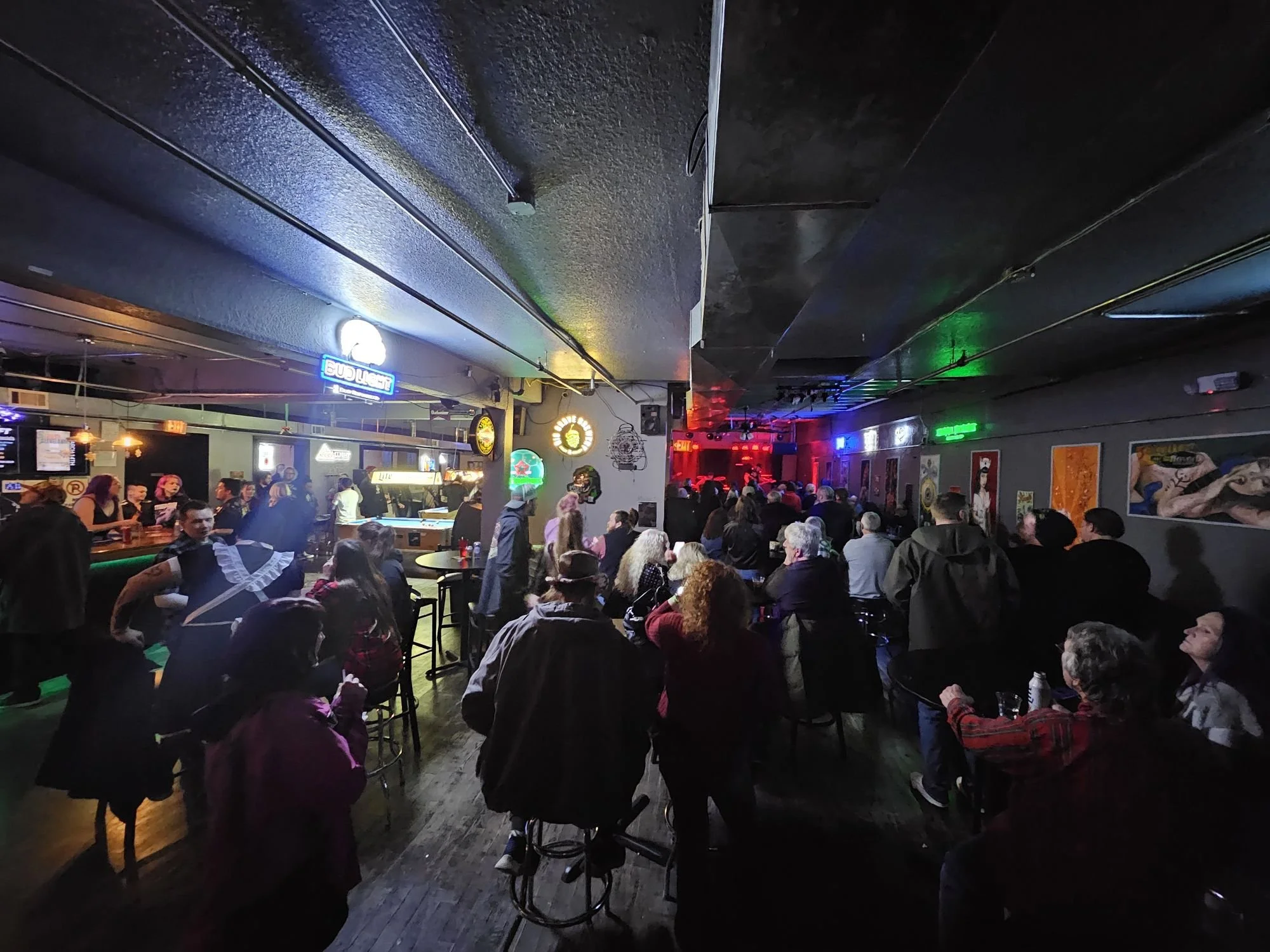 People in a lively bar or nightclub, some sitting and some standing, with neon signs and artwork on the walls, and a stage or performance area at the back.