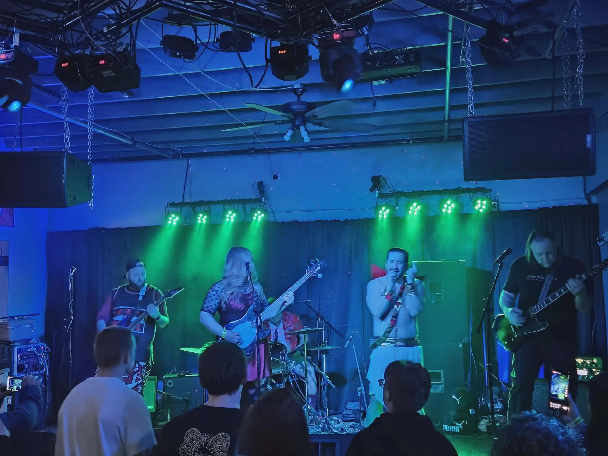 A group of musicians performing on stage with green lighting, musical instruments, and sound equipment, surrounded by an audience in a dark indoor venue.