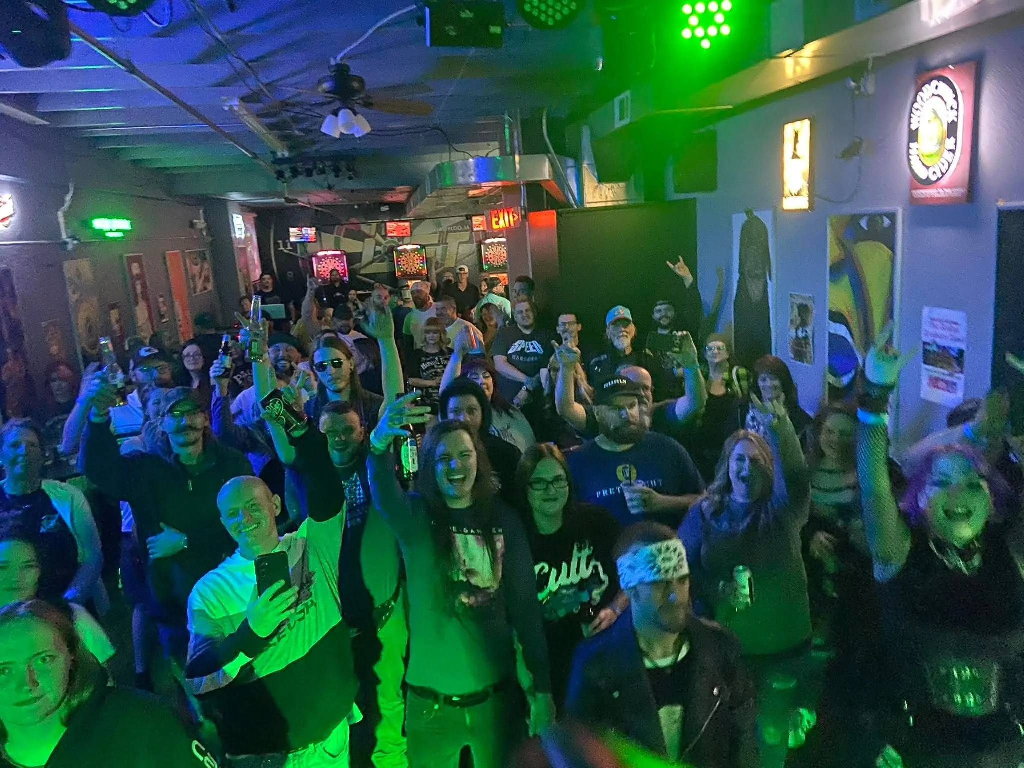 People gathered in a dark room with colorful lights, some holding drinks and taking selfies, standing in front of dartboards and wall decorations.
