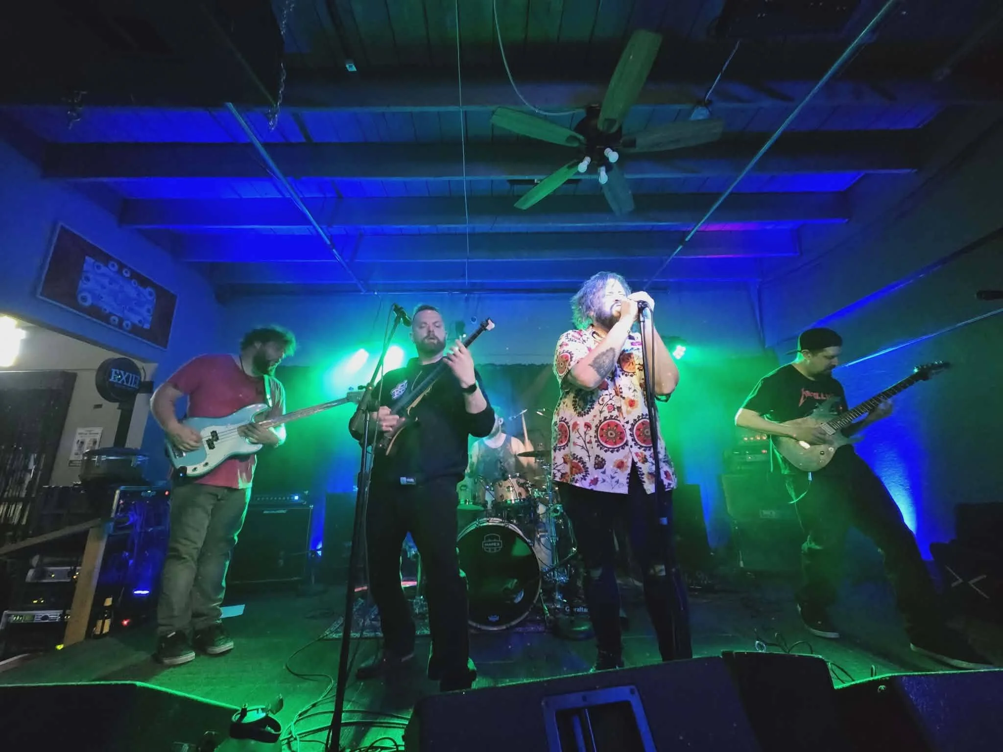 A band performing live on stage with colorful lighting, including a singer in a patterned shirt, guitarists, and a drummer.