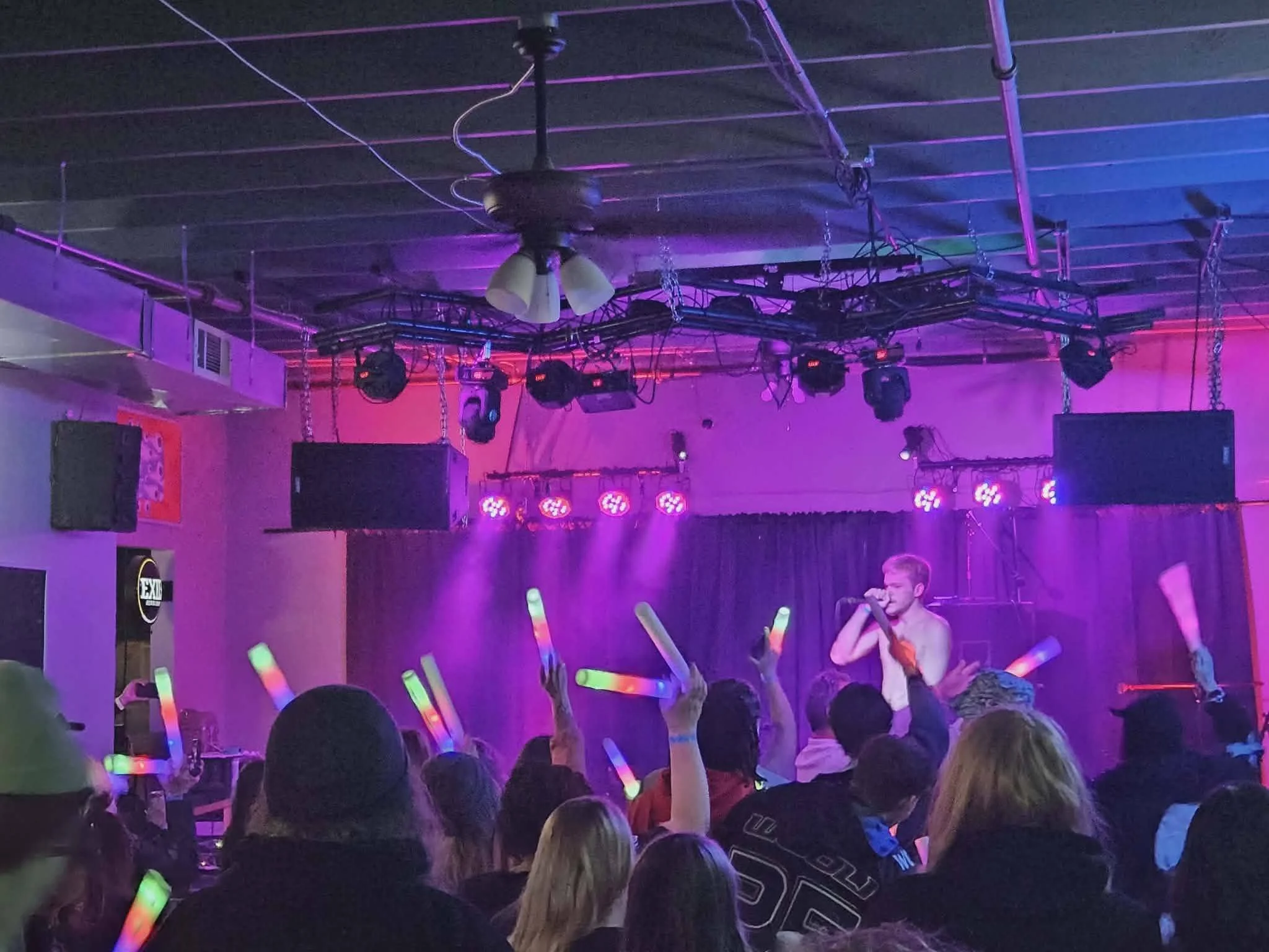 A lively indoor concert or karaoke event with a crowd enjoying glow sticks, illuminated by purple and pink stage lighting, with a performer singing on stage.