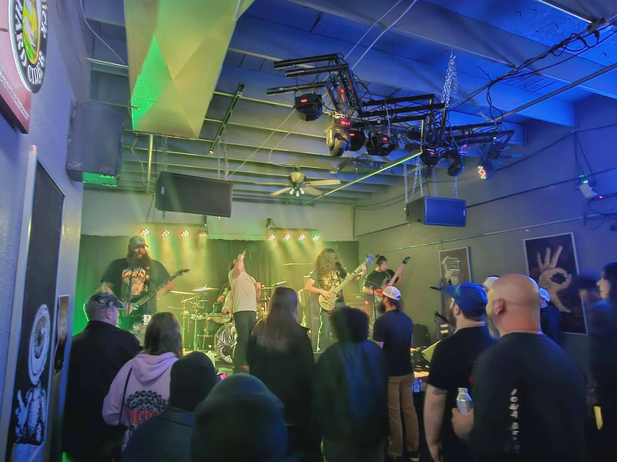 A band performs on a stage with green and yellow lighting, featuring guitarists and a drummer, at a live concert with an audience watching.
