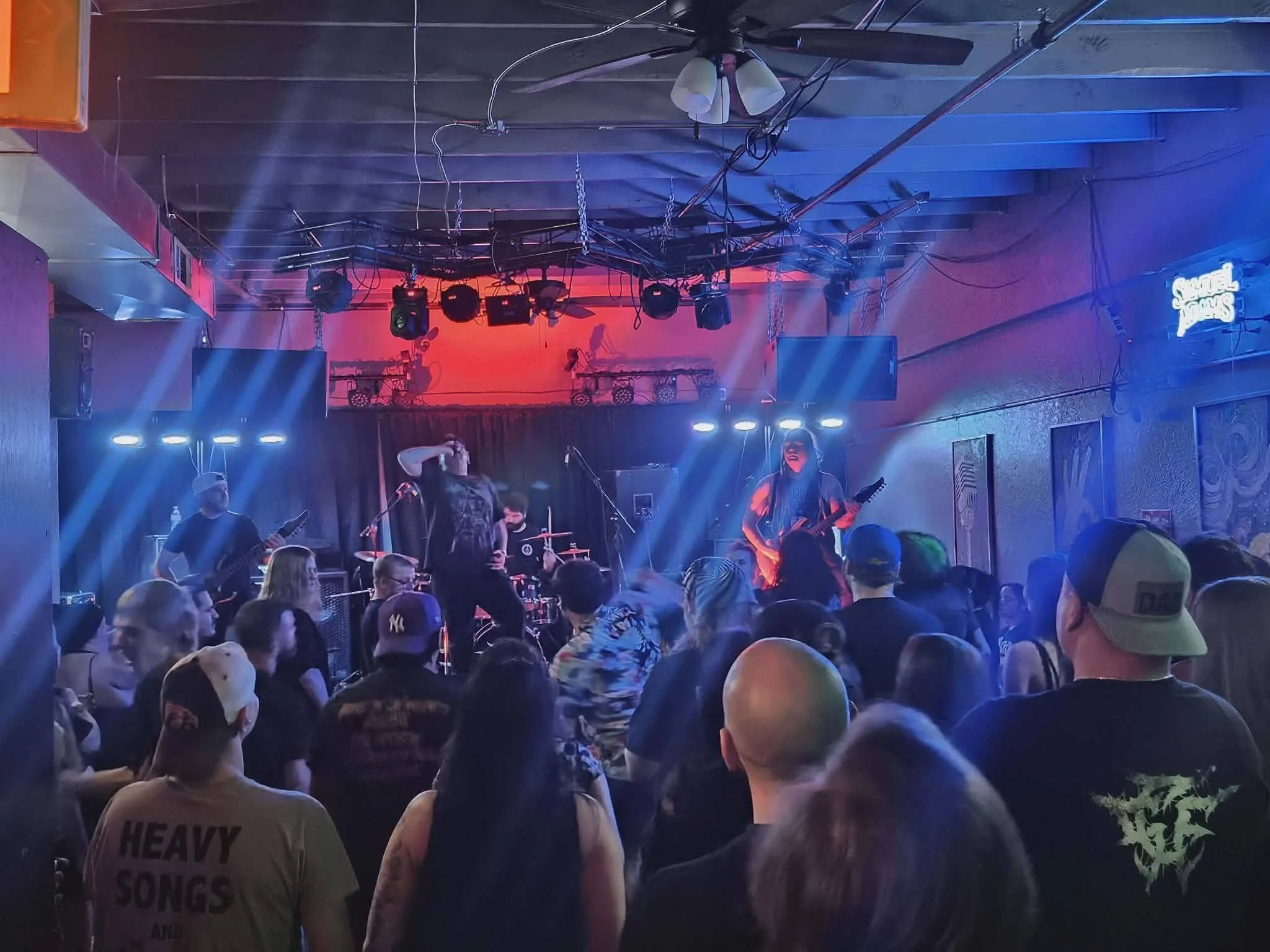 A group of people attending a live music performance in a dimly lit indoor venue with stage lighting. The audience is watching the musicians on stage, with some wearing hats, including military caps. The stage is equipped with musical instruments, mi