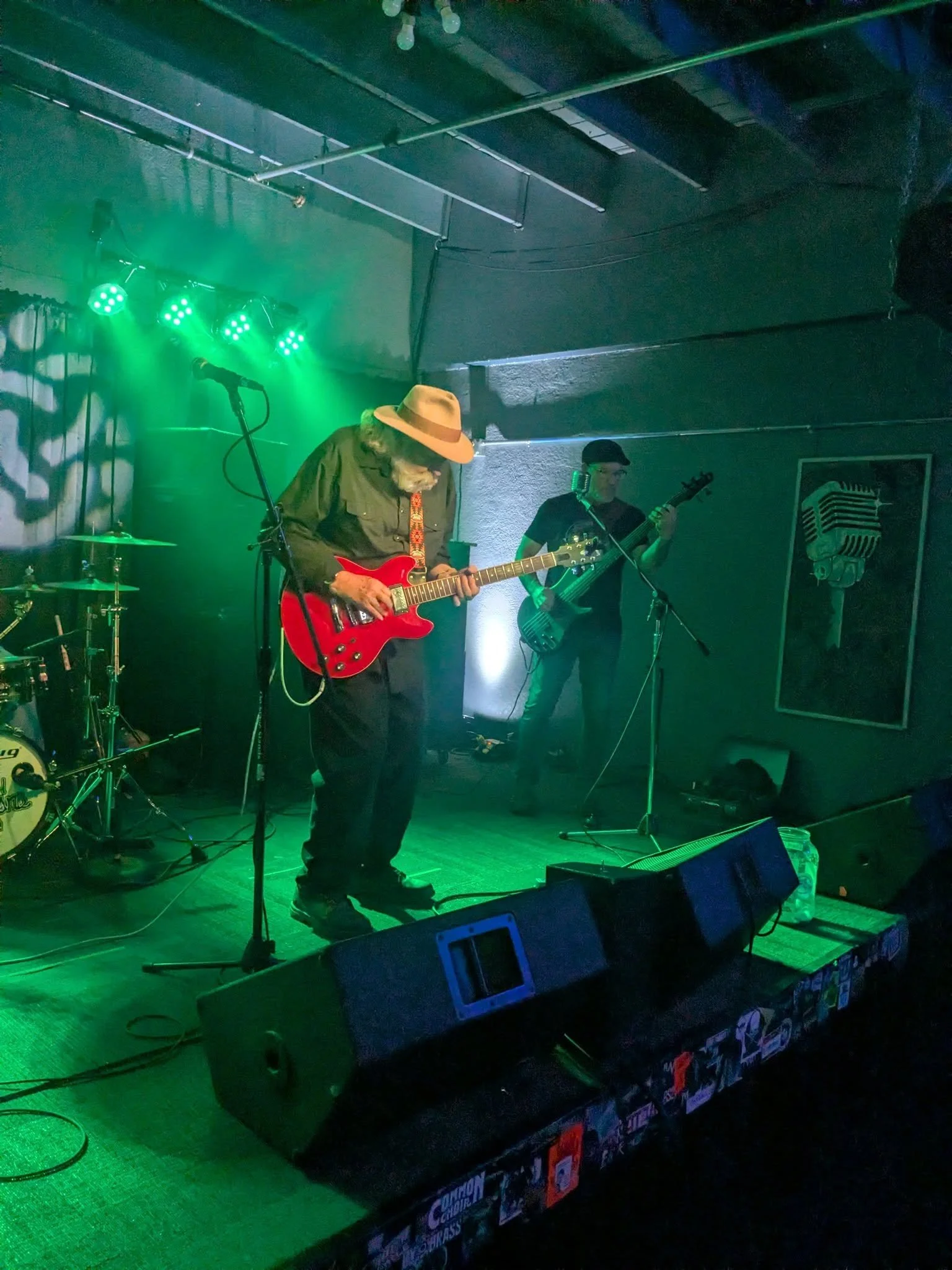 Musicians performing on stage with green lighting, one playing an electric guitar, and another on bass guitar, in an indoor venue.