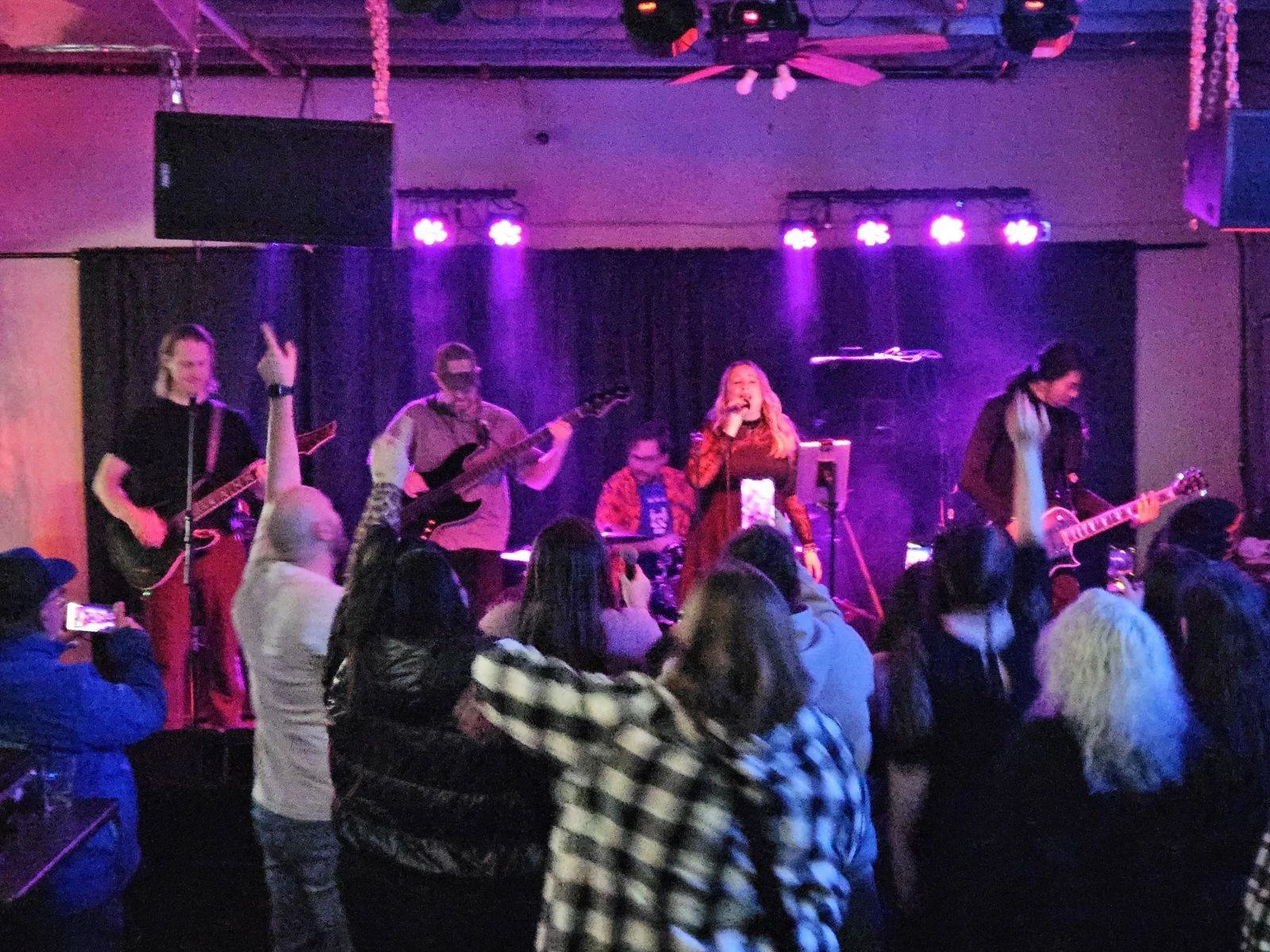 Band performing on stage with purple lighting, audience watching, some taking photos.