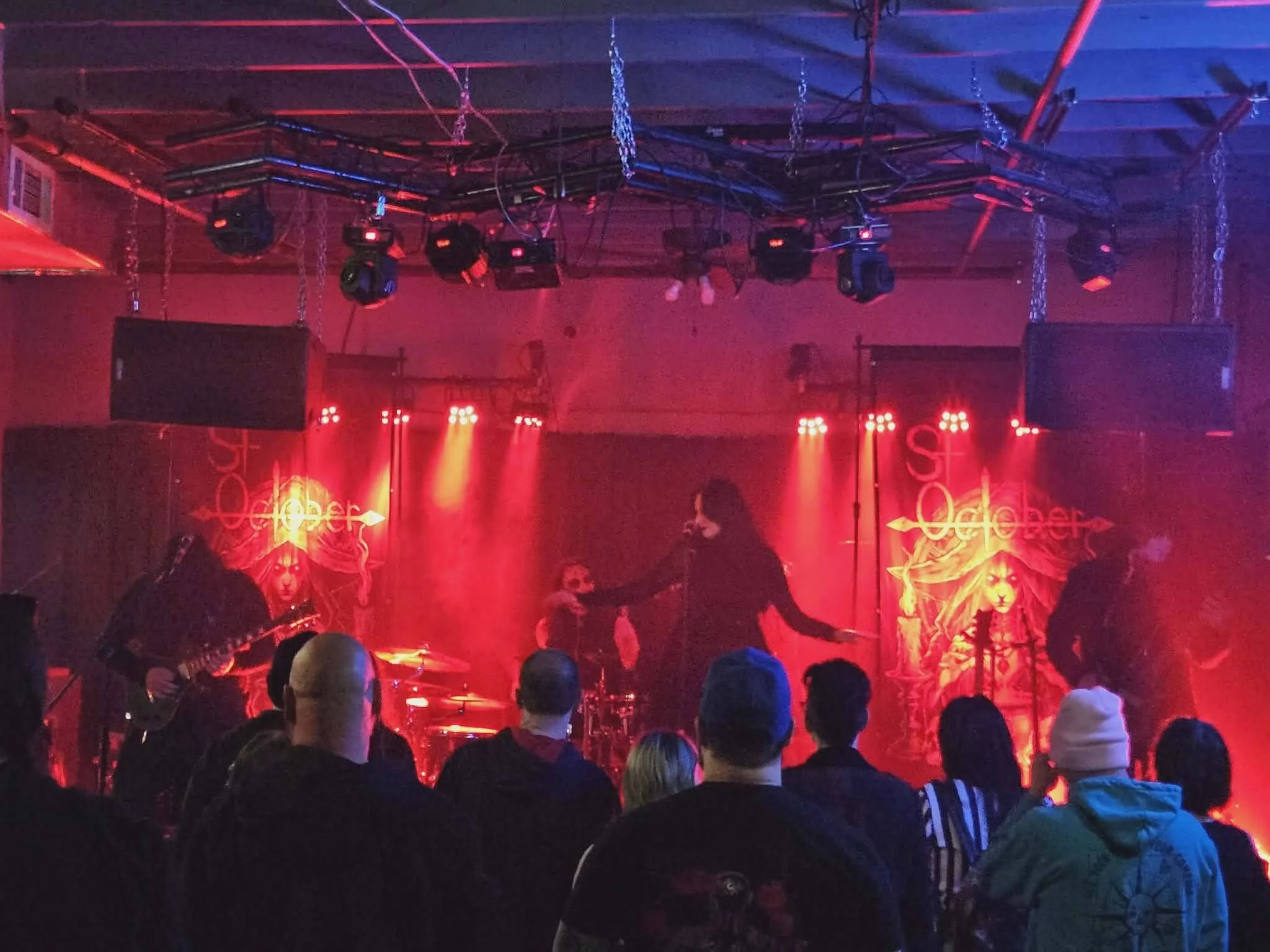 A group of people are gathered at a concert or live music event with a stage set up, featuring red and orange lighting, silhouettes of performers, and a dark atmosphere.