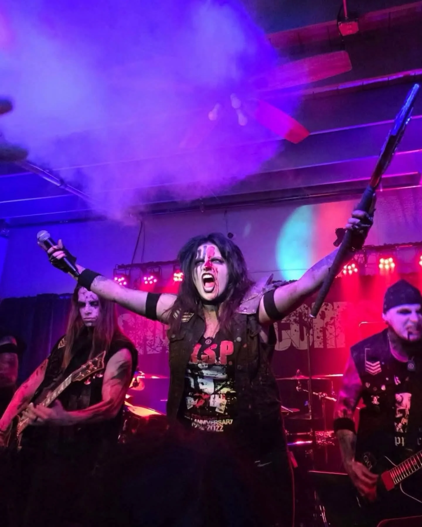 A band performing on stage with purple and red stage lighting, a woman with dark hair in the center holding a microphone and raising her arms, and other musicians playing guitars beside her.
