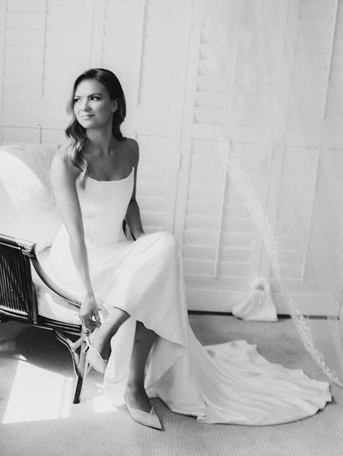 Black and white photo of a woman sitting on a chair in a white dress, looking to the side with a slight smile.