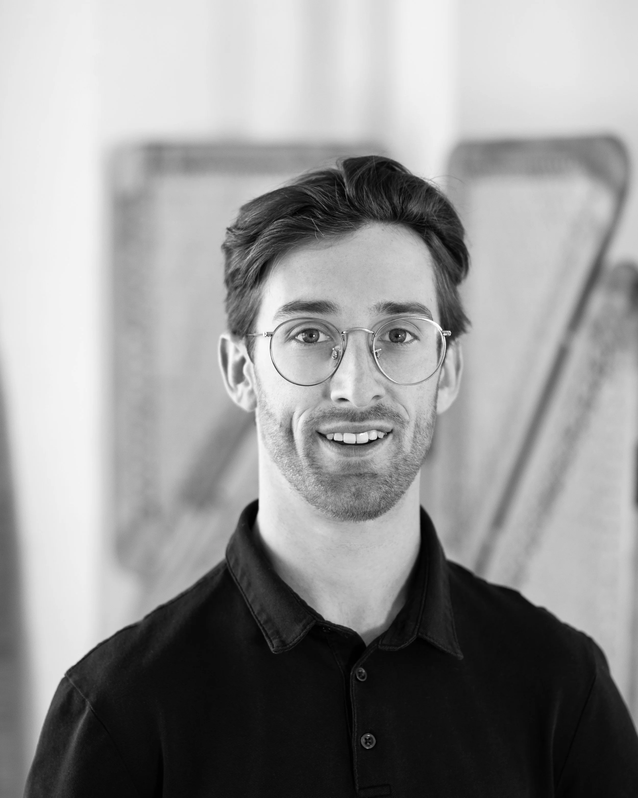 Black and white portrait of a smiling young man with glasses, wearing a dark collared shirt, in front of a blurred background.