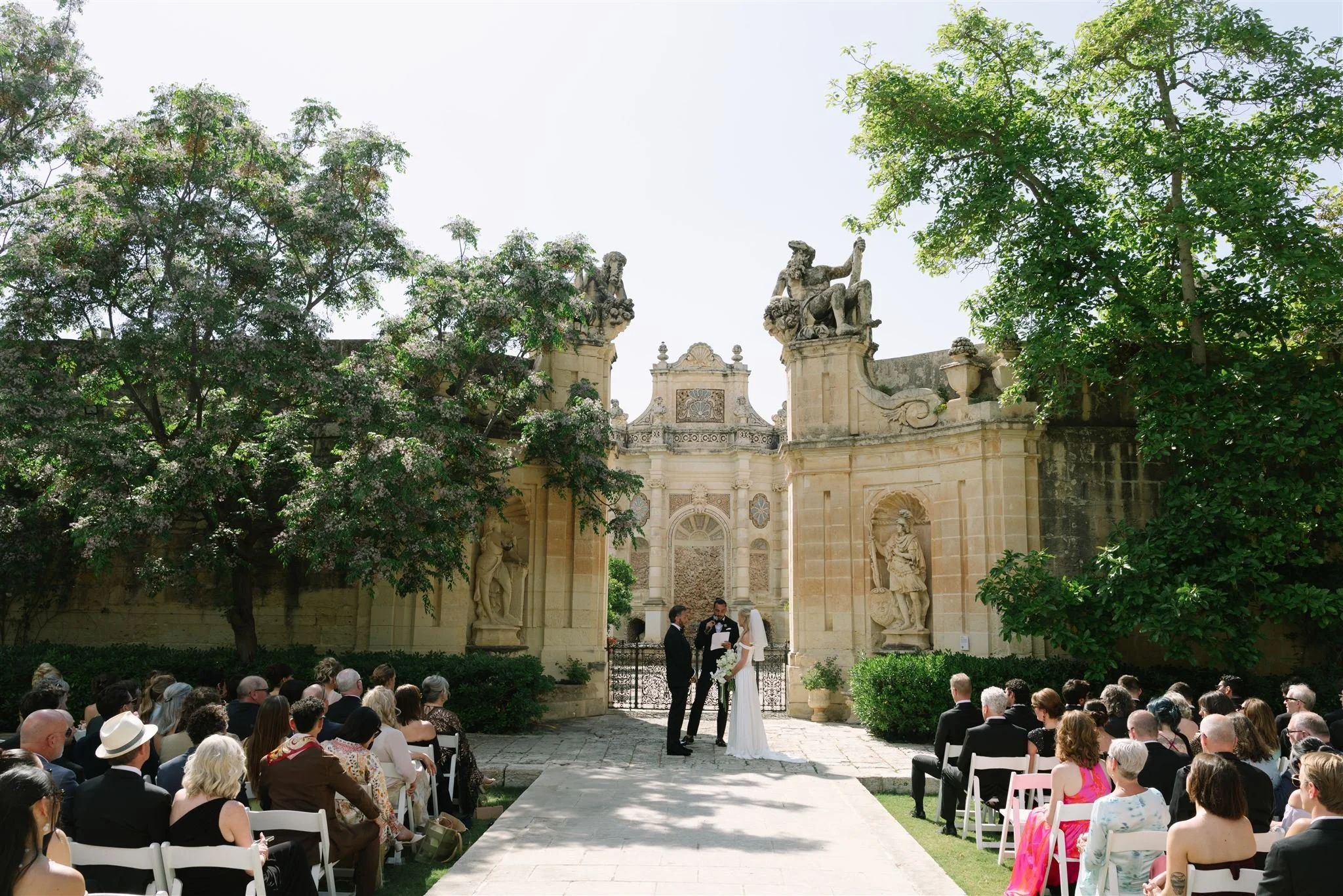 Wedding ceremony outdoors at a historic stone mansion with statues and lush green trees, guests seated on white chairs, bride and groom standing in front of officiant.