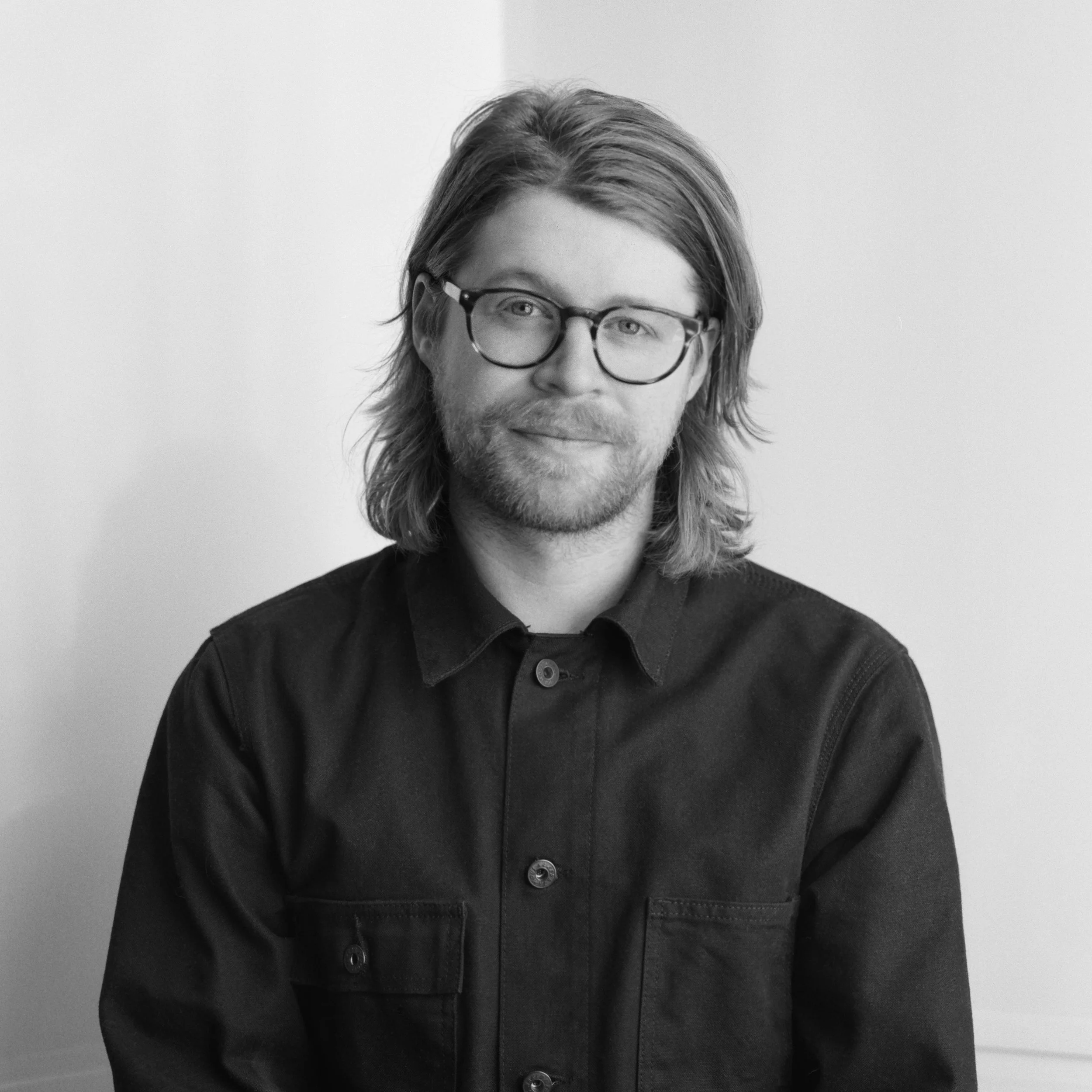 Black and white portrait of a man with shoulder-length wavy hair, glasses, and a beard, wearing a dark button-up shirt, smiling slightly and looking at the camera.