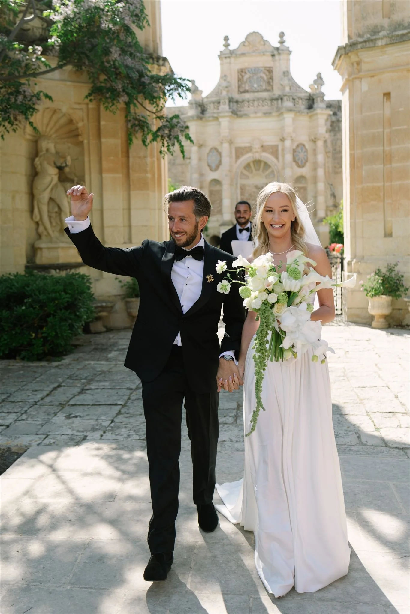 Happy newlywed couple walking out of a historic building with a stone facade, holding hands, and celebrating their wedding day. The groom is wearing a black tuxedo and bow tie, and the bride is in a white gown holding a large bouquet of white flowers. An officiant or groom is visible in the background.