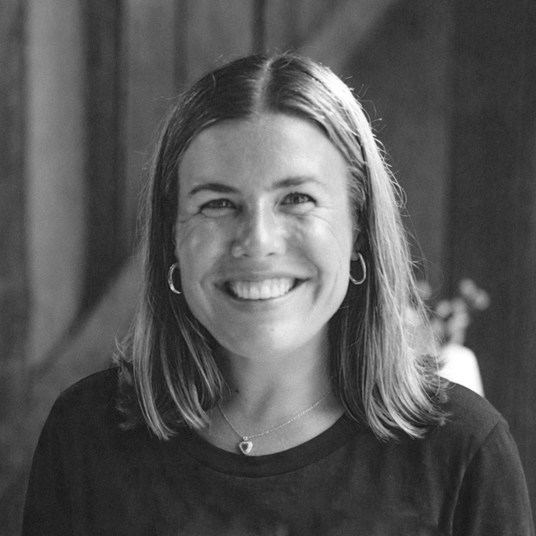 Black and white photo of a smiling woman with shoulder-length hair, wearing earrings and a necklace, indoors with a wooden wall in the background.