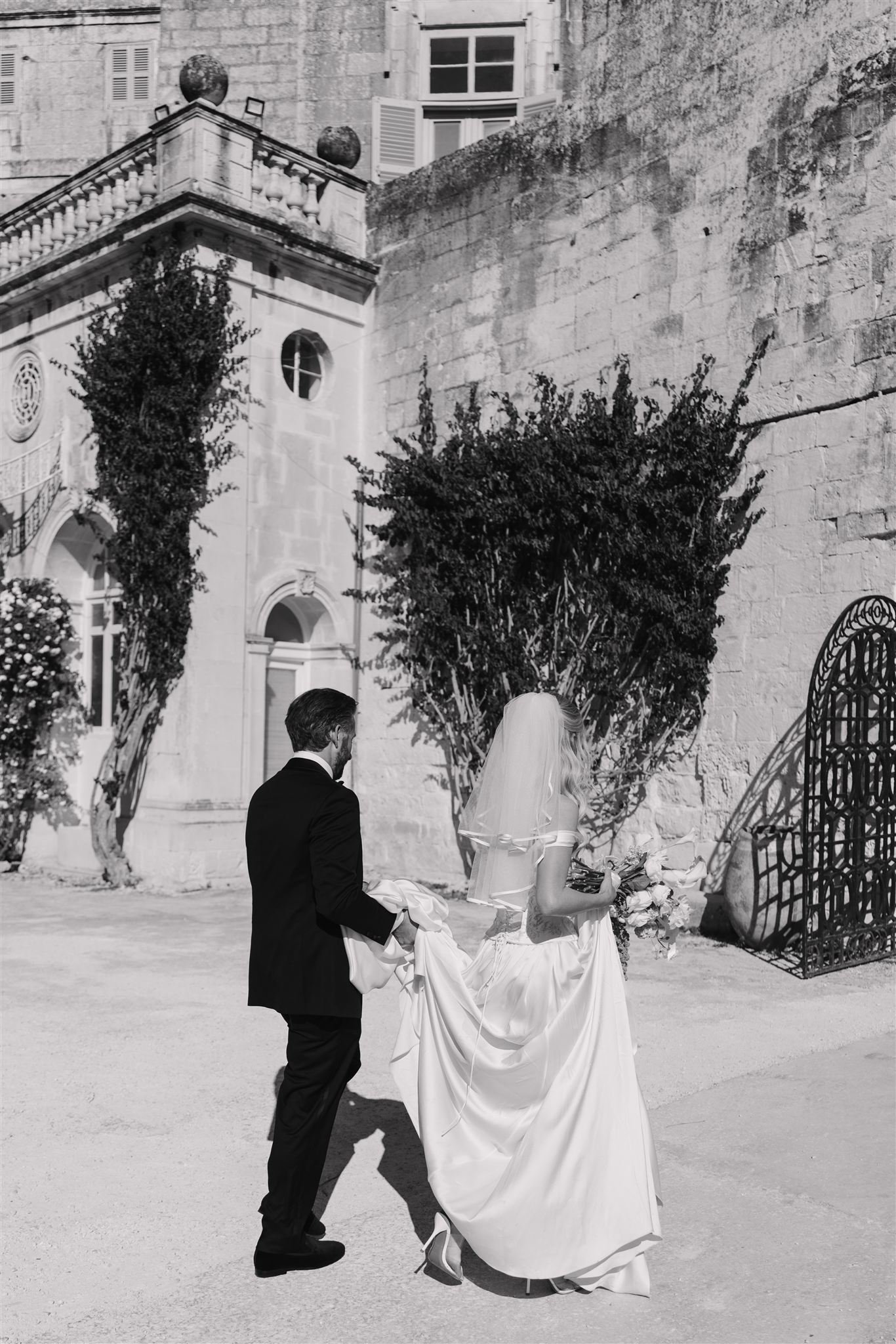 A black and white photo of a bride in a wedding dress holding a bouquet, walking beside a man in a suit, outdoors near a stone building with trees and a metal gate.
