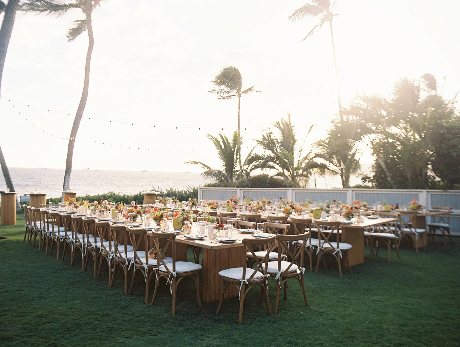 Outdoor dining setup with long wooden tables and chairs, decorated with colorful flower arrangements, set on green grass near the coast with palm trees and string lights, during sunset.