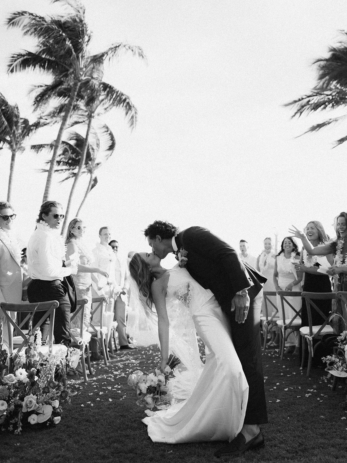 A black and white photo of a wedding ceremony outdoors with palm trees. A couple is kissing, the groom dipping the bride as she holds a bouquet, while guests look on and smile.