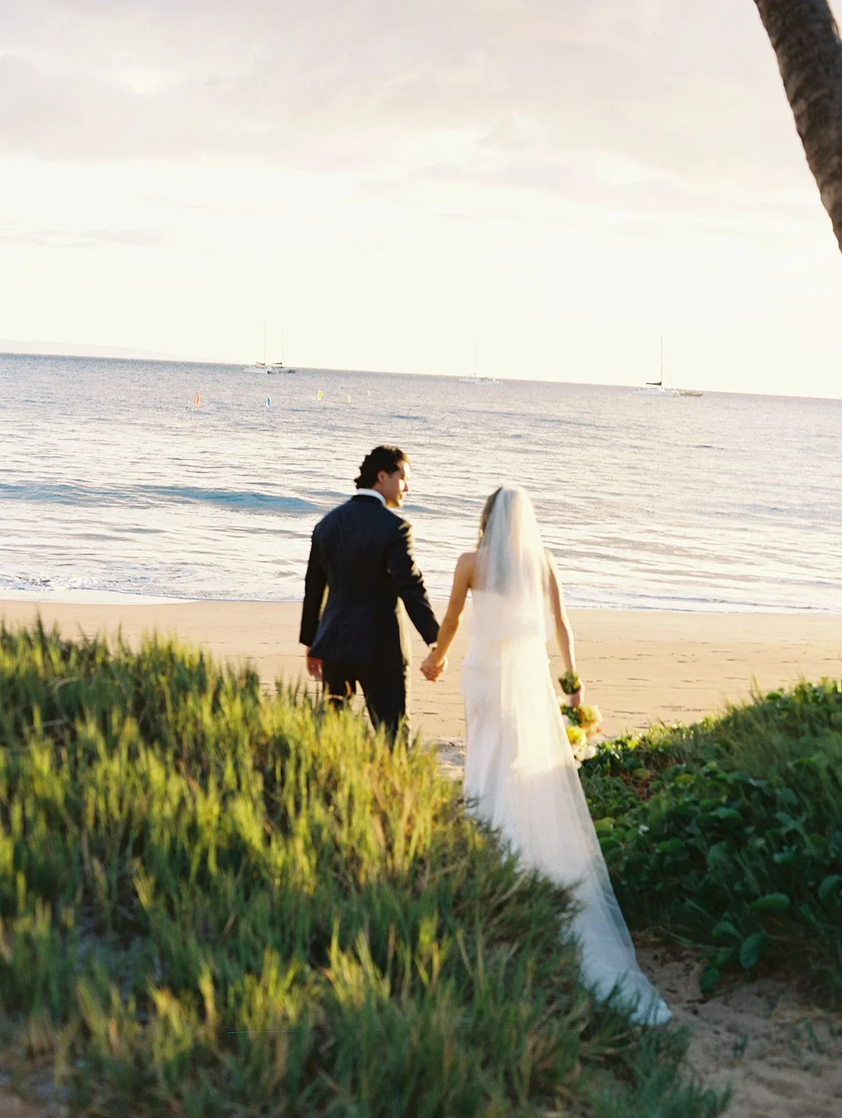 A bride and groom walking hand in hand on the beach near the water, with sailboats in the distance, during sunset.
