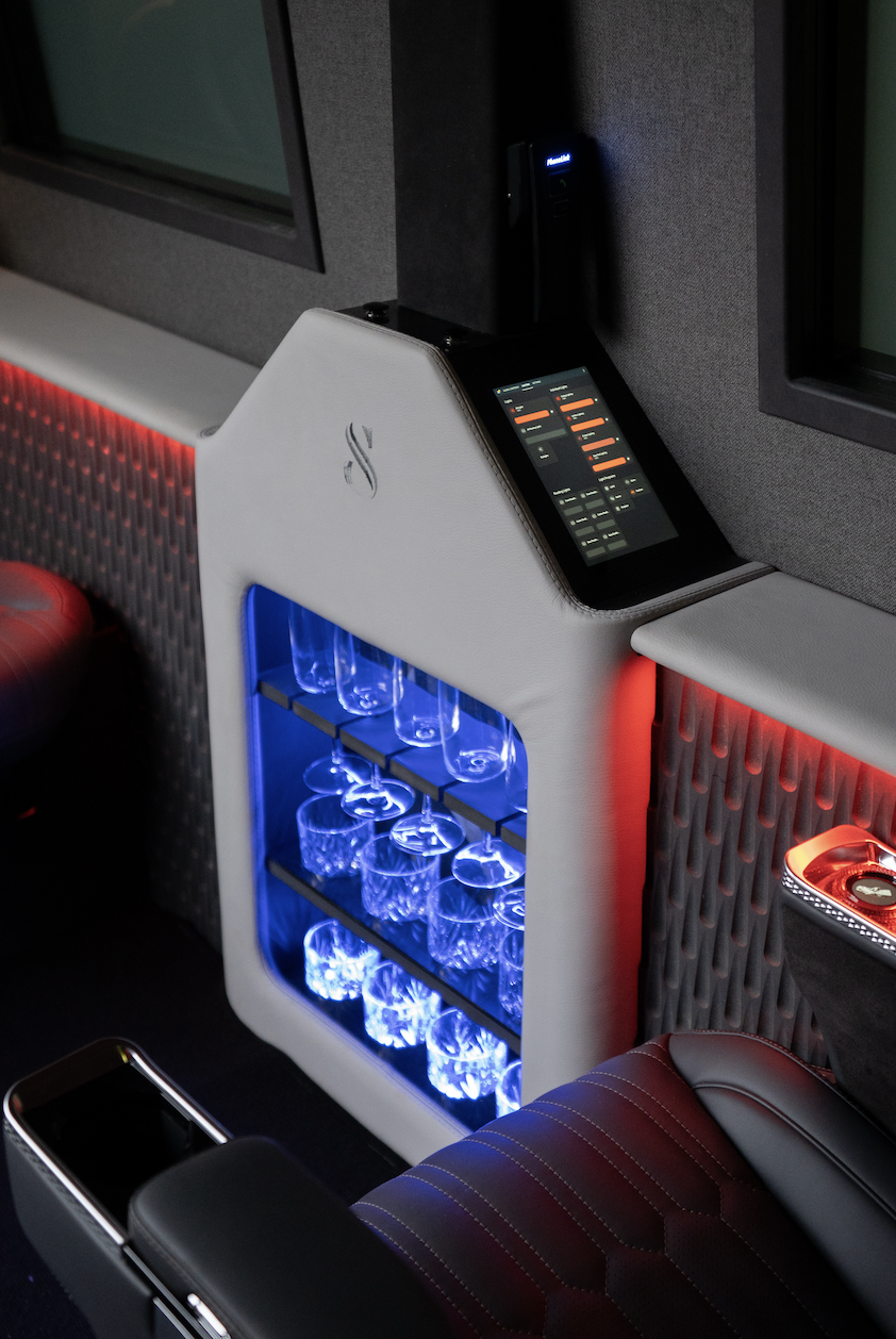 A modern built-in bar area with illuminated glassware shelf, a touchscreen control panel, and leather seating in a luxury vehicle or lounge.