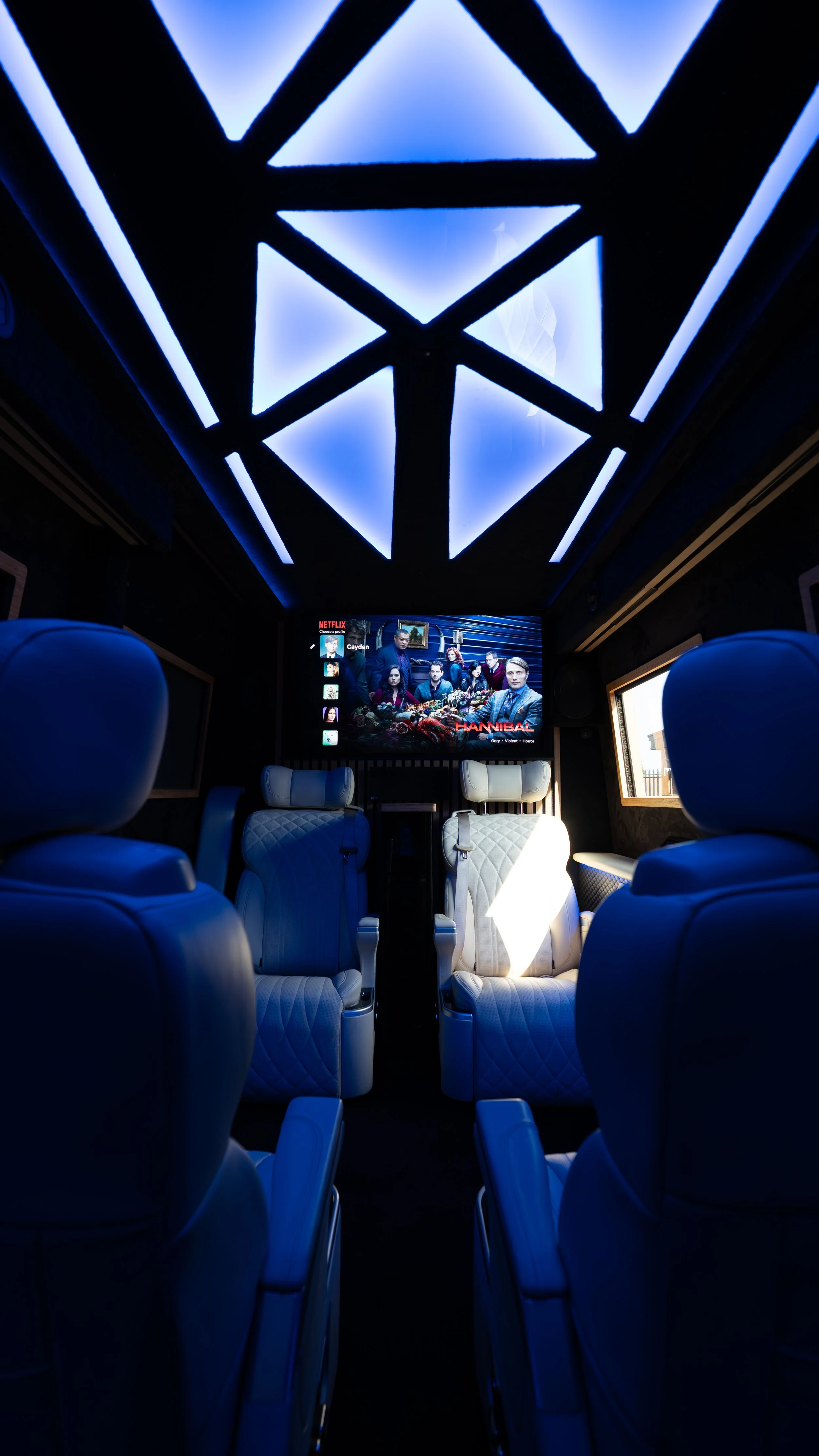 Luxury home theater room with six white leather recliner seats, a large screen displaying the show 'Hannibal,' and blue neon ceiling lights.