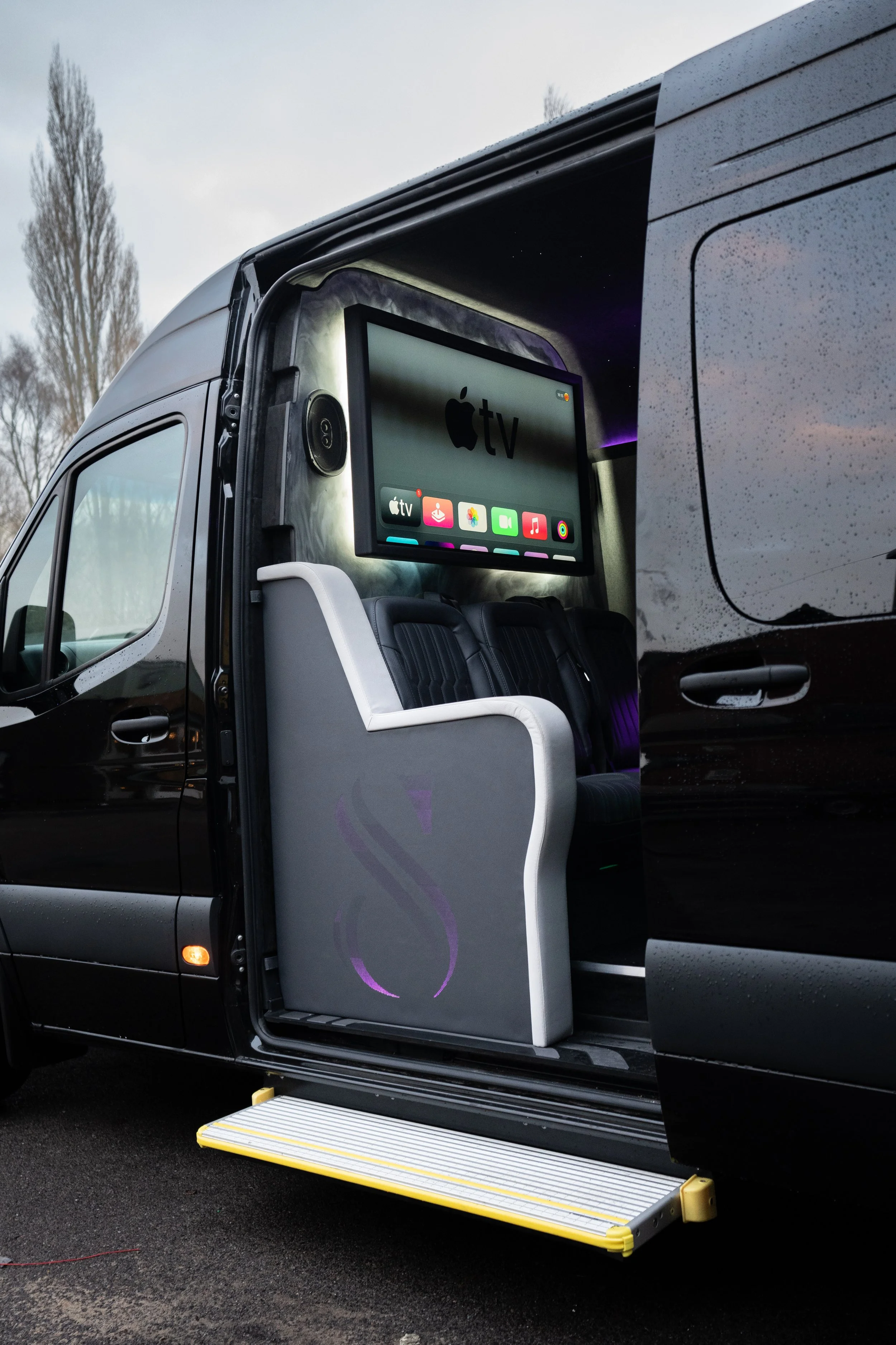 Inside a black van with an open side door, featuring a large screen displaying the Apple TV interface, with seats and a step at the entrance.