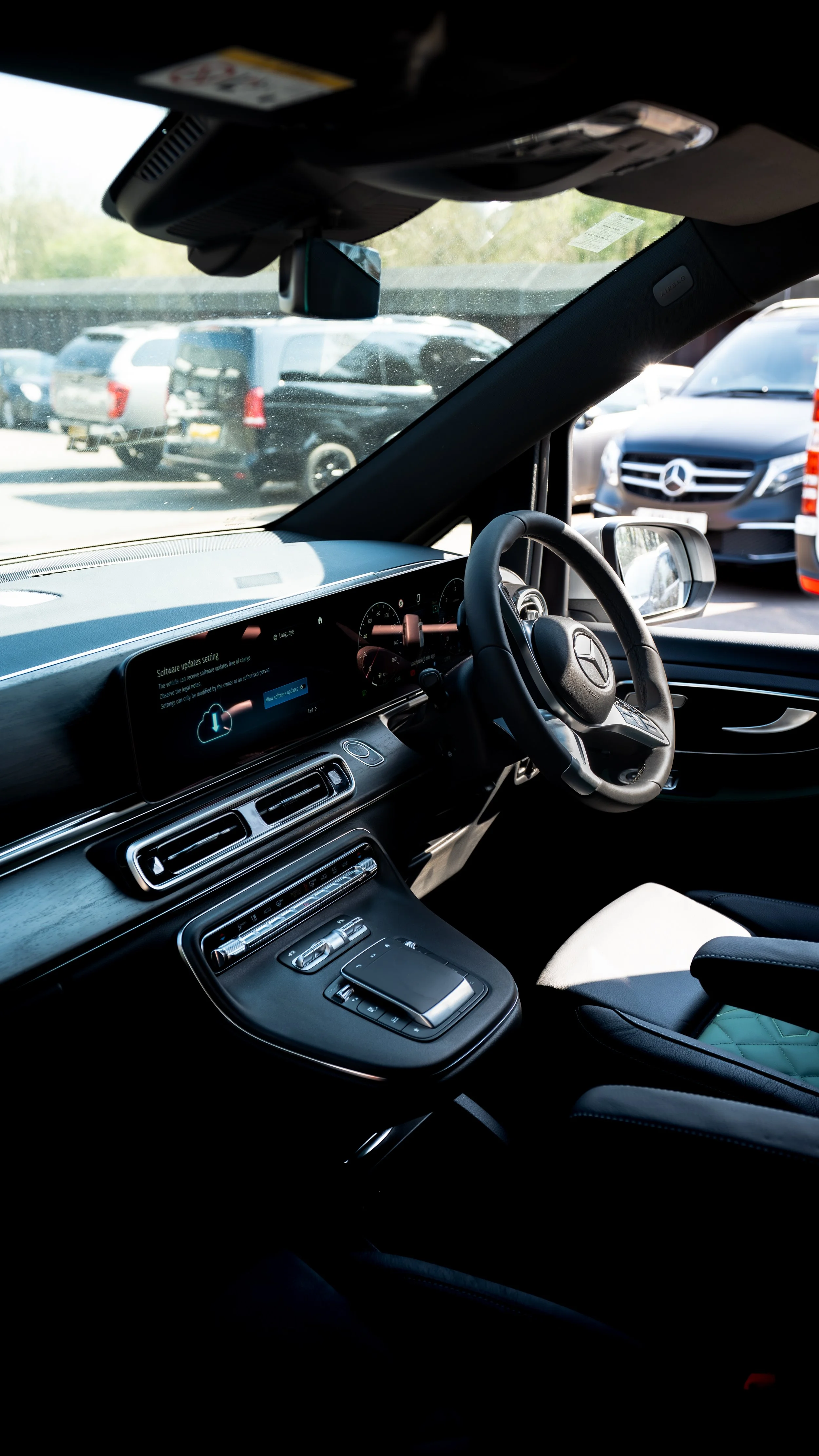 Interior of a modern Mercedes-Benz vehicle, showing the dashboard, steering wheel, and central console with controls, parked in a lot with other cars visible through the windshield.