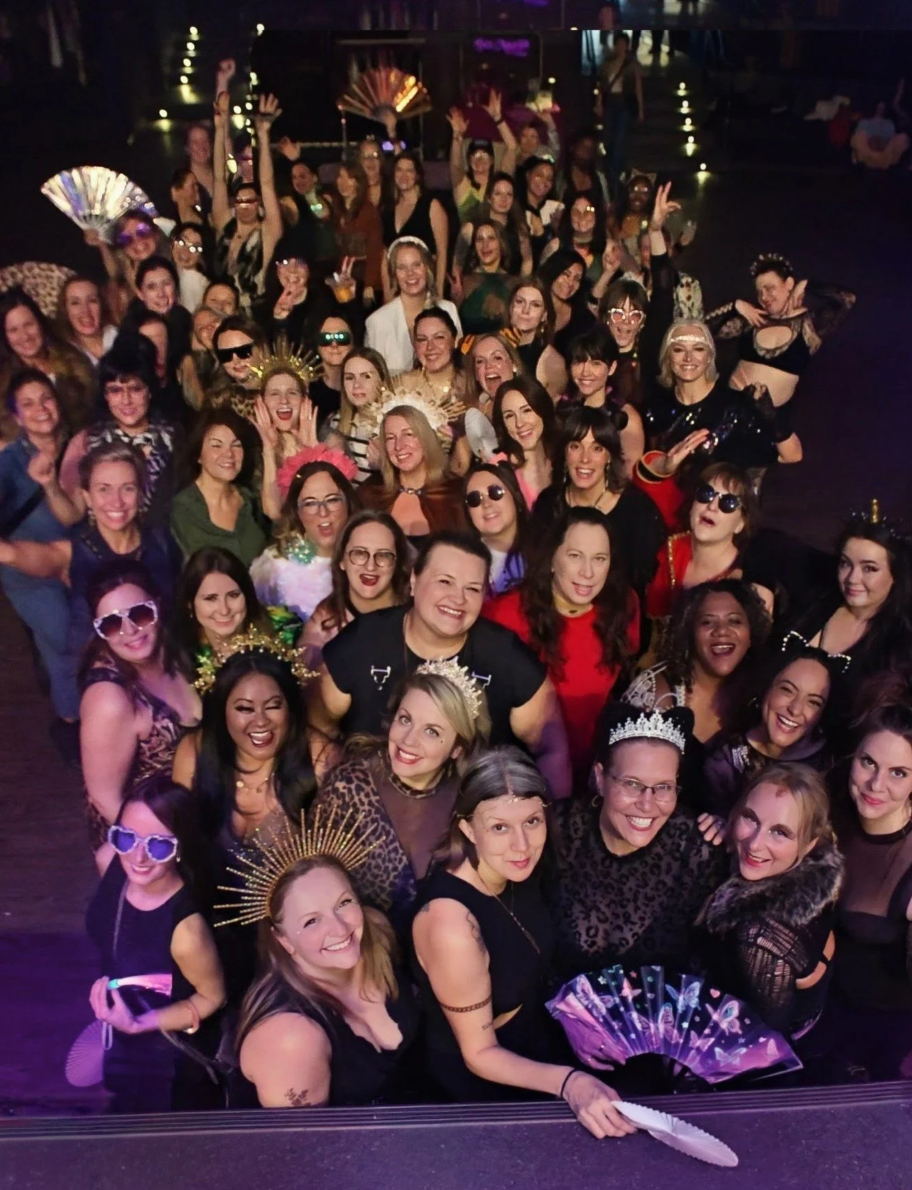 Group of women at a celebration or party, some wearing crowns, tiaras, and fun accessories, posing for a group photo in a dimly lit venue.
