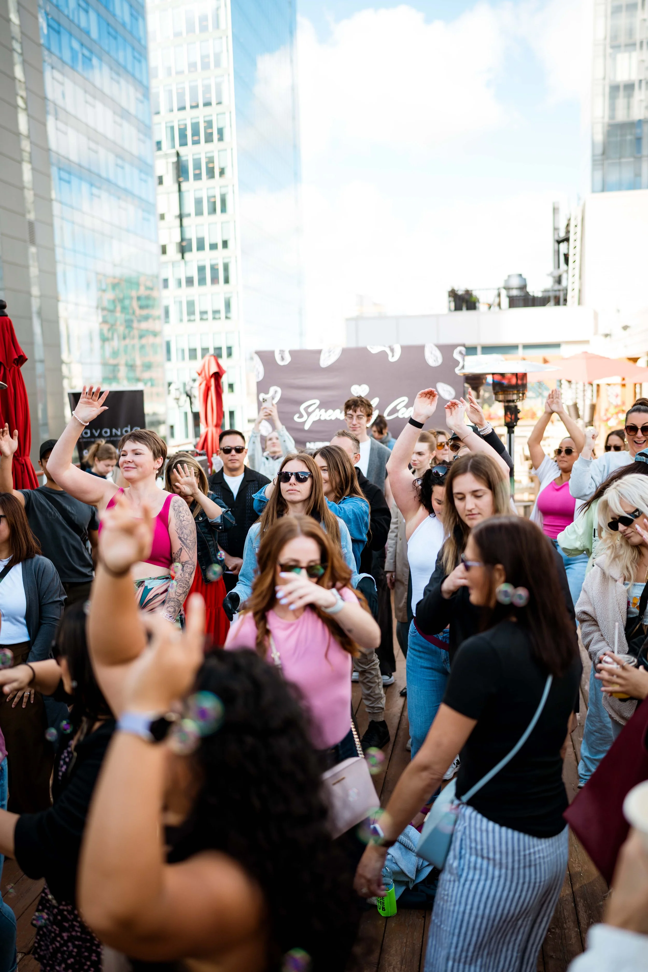 A crowd of people gathered outdoors on a sunny day, some dancing and raising their hands, with tall modern buildings and a banner in the background.
