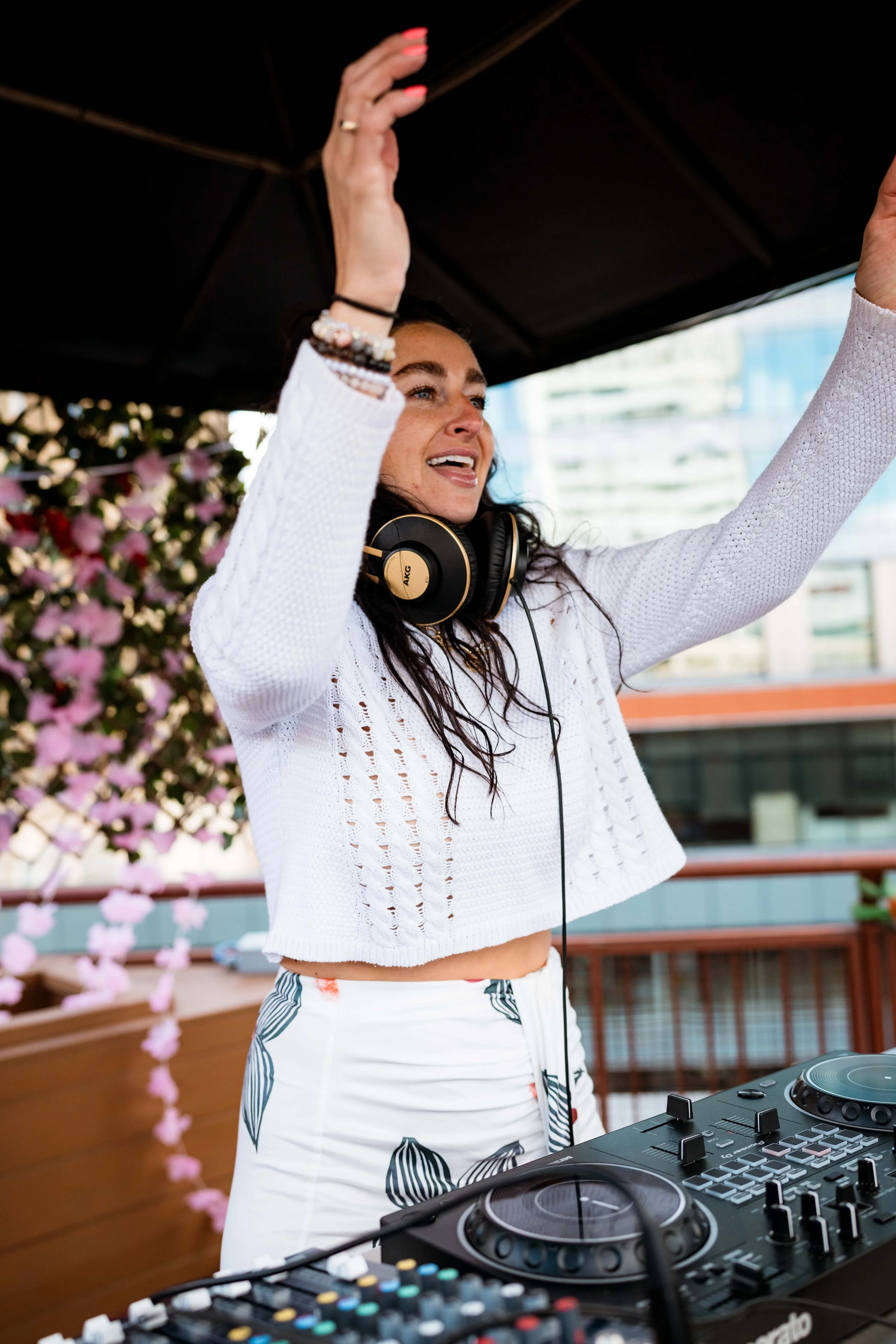 A female DJ with headphones is at a turntable, raising her hands in the air, smiling, with pink and purple flowers and modern glass buildings in the background.