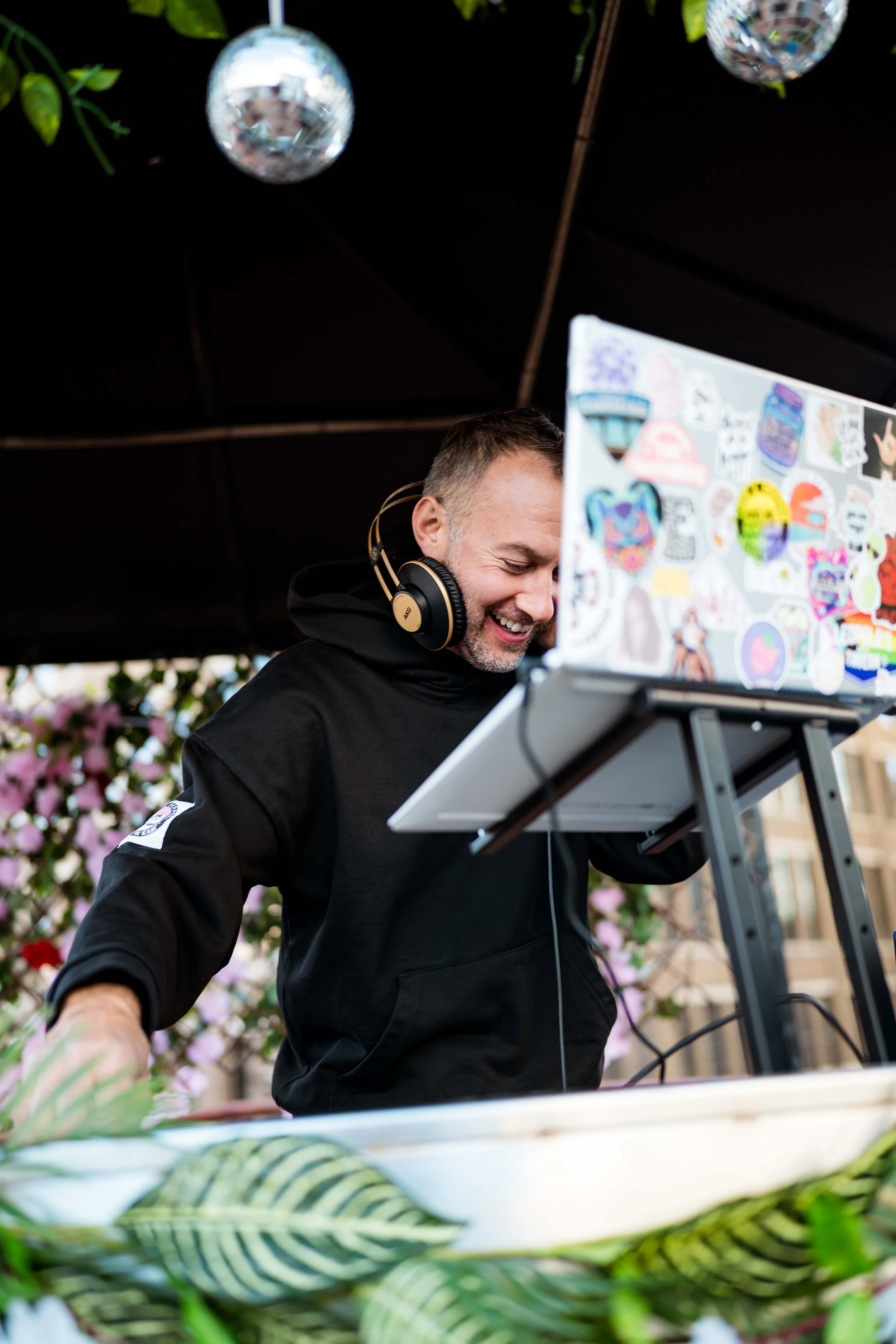 A man wearing a black hoodie and headphones is DJing at an outdoor event, smiling while standing behind a laptop decorated with colorful stickers. Suspended disco balls are hanging overhead, and pink flowering plants are visible in the background.