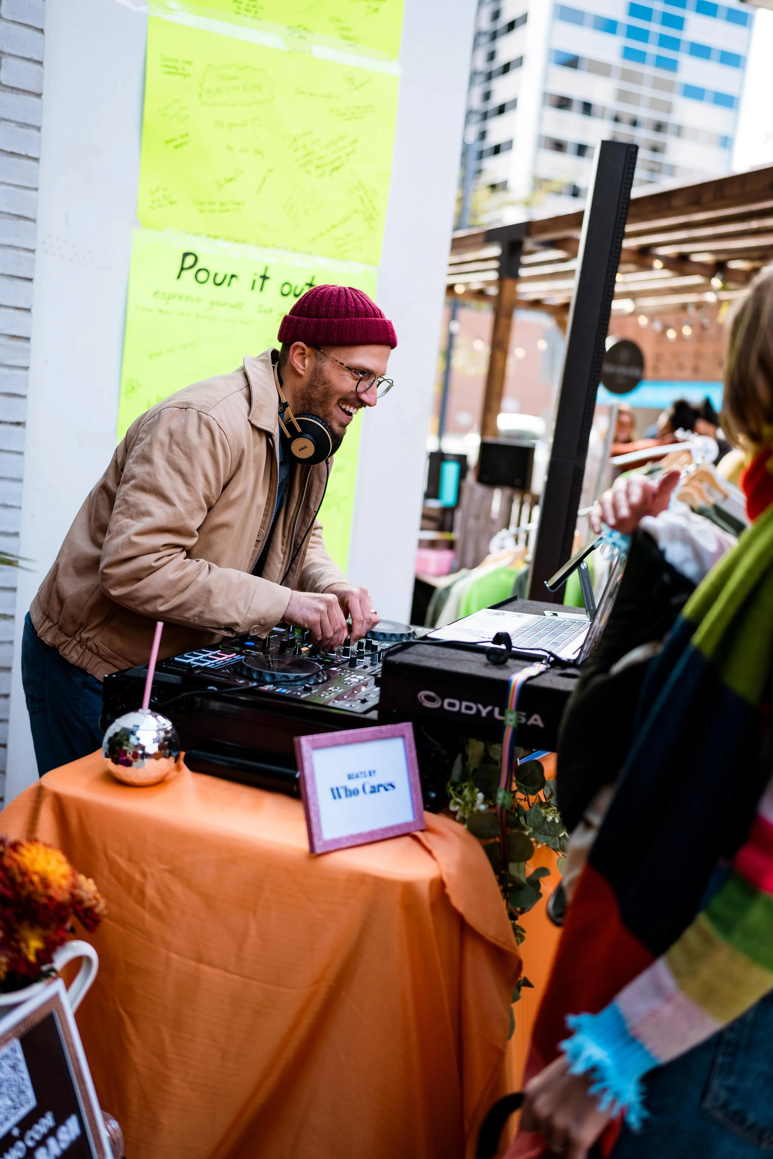 A man with glasses and a red beanie DJing at an outdoor event, smiling, while a person in front watches. The DJ table has a sign that says 'SAFE SPACE Who Cares'. There are notes posted on a wall behind him.