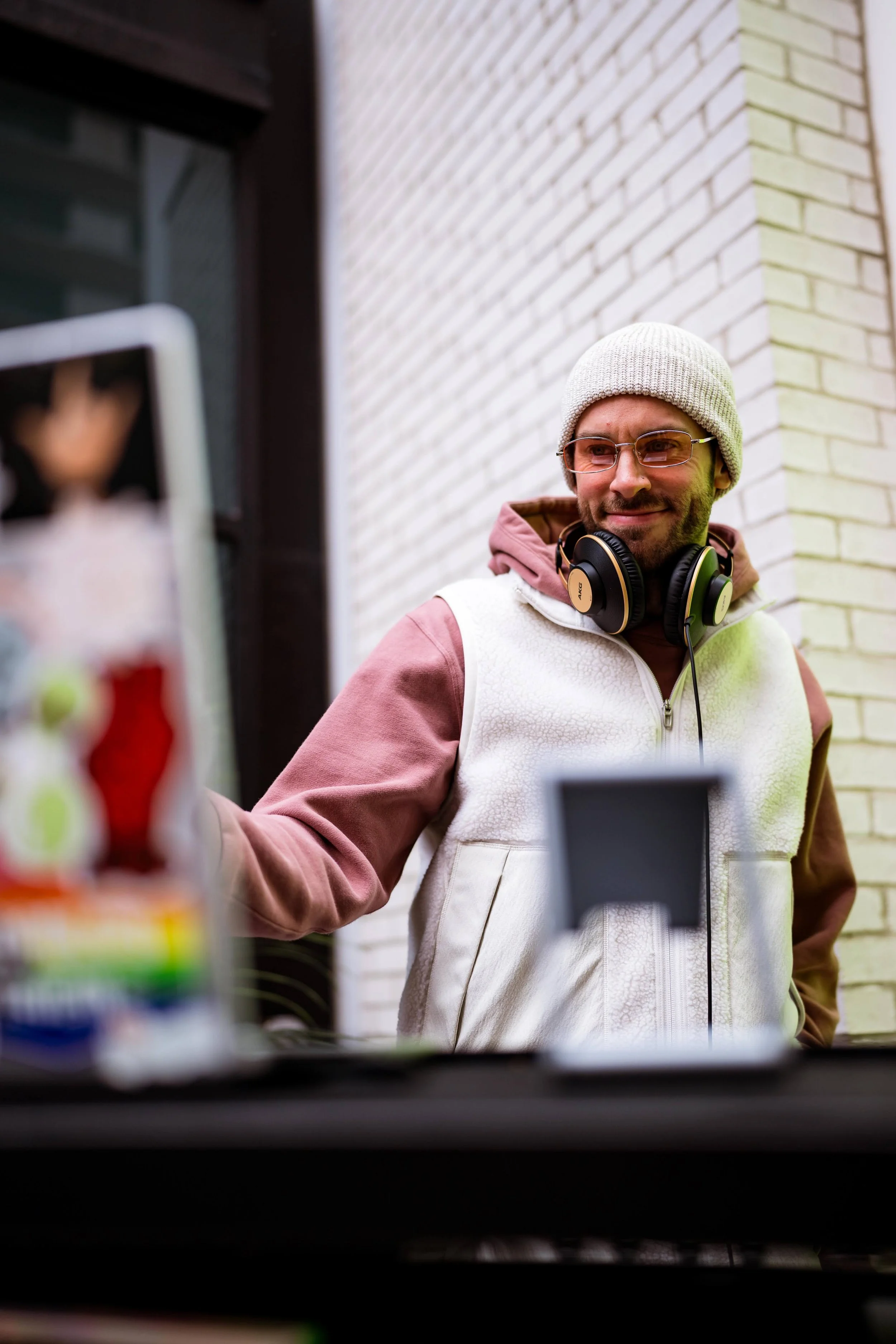 A man with glasses, wearing a beanie and headphones around his neck, stands outside near a white brick wall, smiling as he interacts with a phone on a stand.