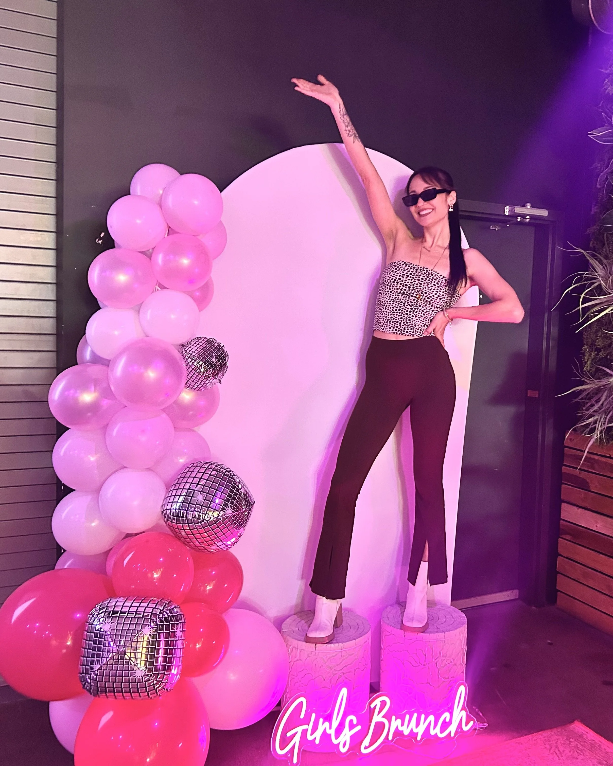 A woman wearing sunglasses, a leopard print crop top, and black pants standing on pink cylindrical platforms with a neon sign that reads 'Girls Brunch.' She is smiling and posing with one arm raised, next to a decorative balloon arrangement with pink, white, and silver disco balls in a dimly lit space.