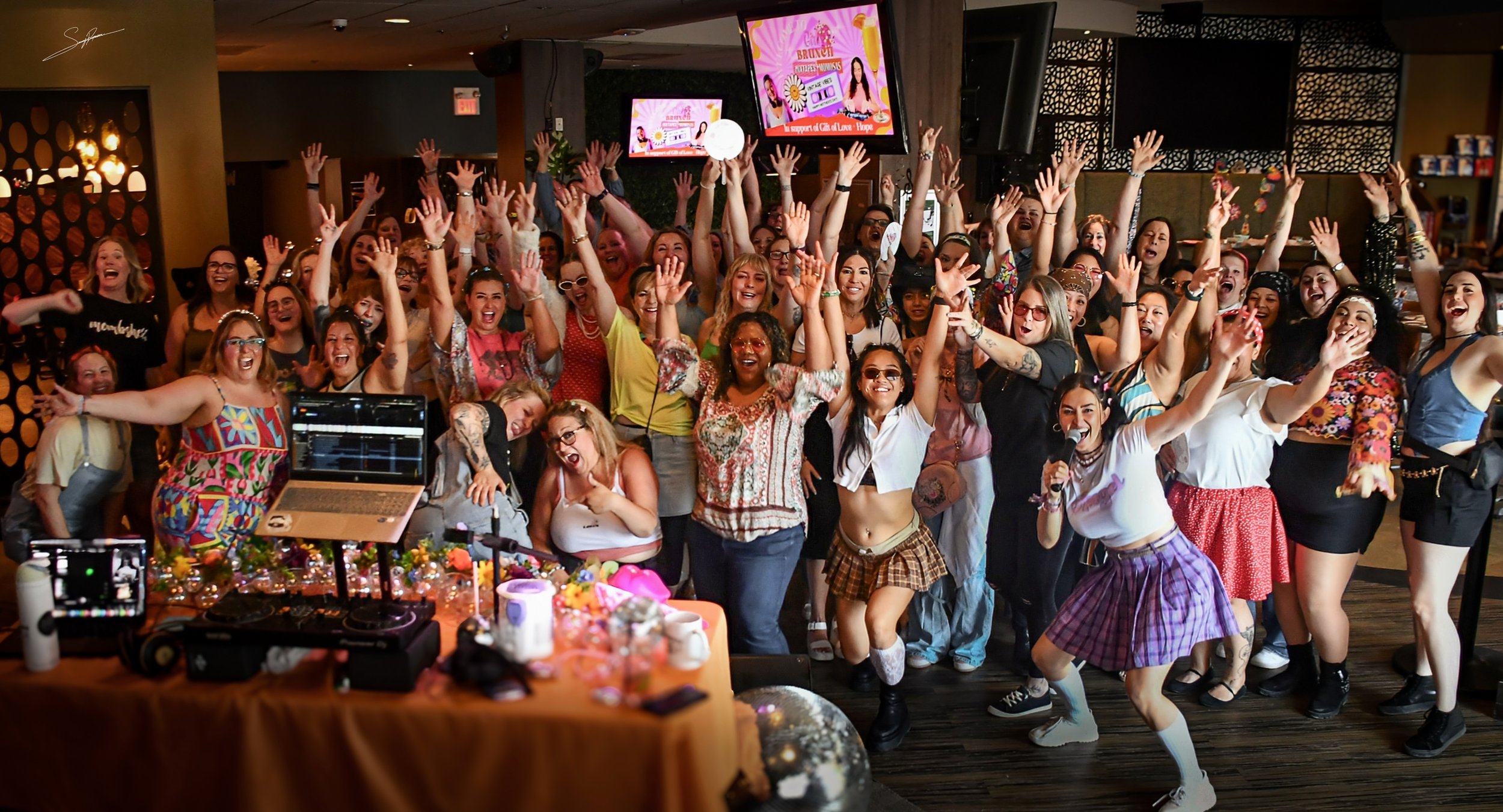 A large group of women at a party dance and cheer in a lively, colorful venue with decorations, DJ equipment, and multiple screens showing party graphics.